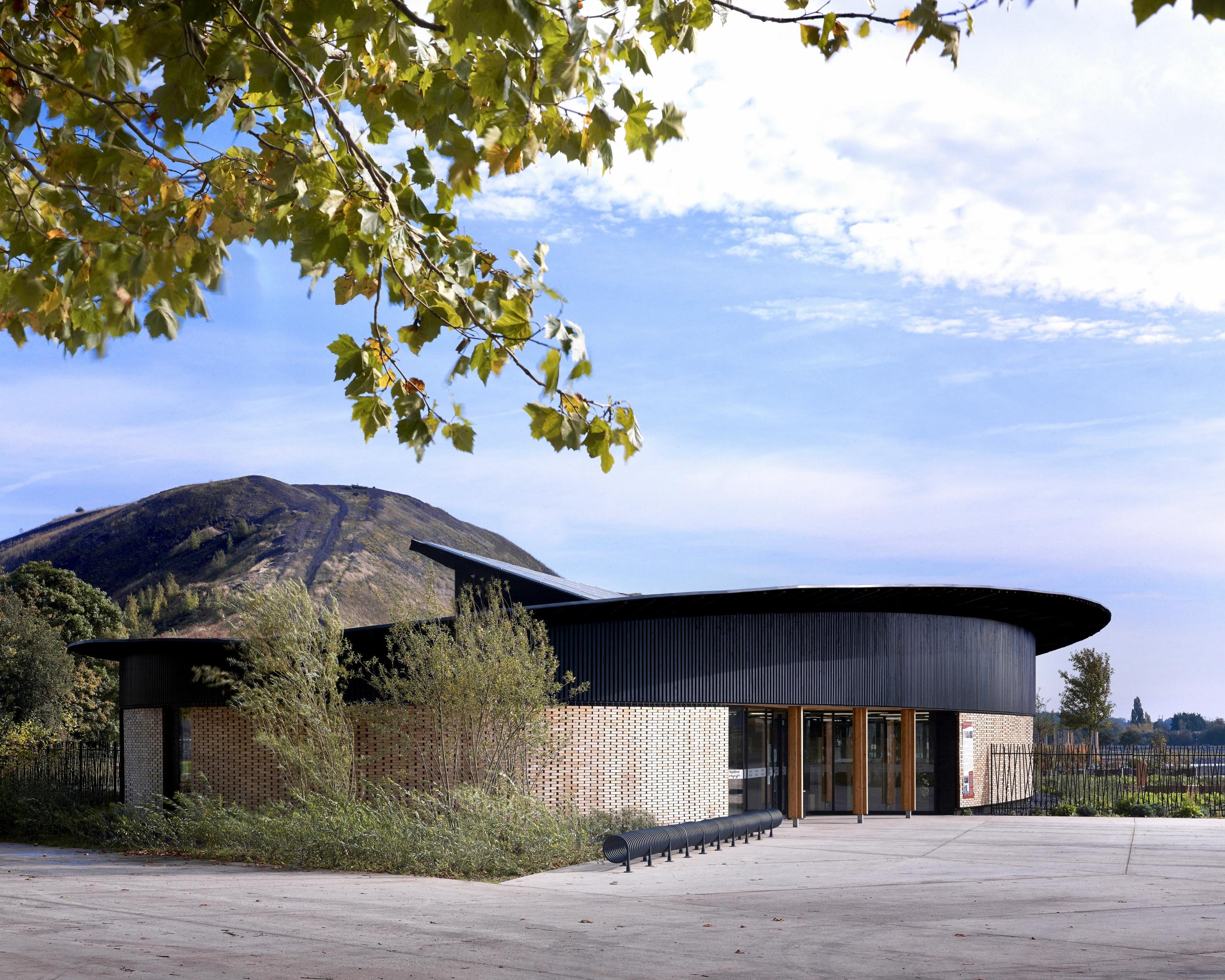 Maison de l’Environnement à Hénin-Beaumont, pavillon ovale en ossature bois et briques de bois, bardage noir, large auvent circulaire face au paysage minier.