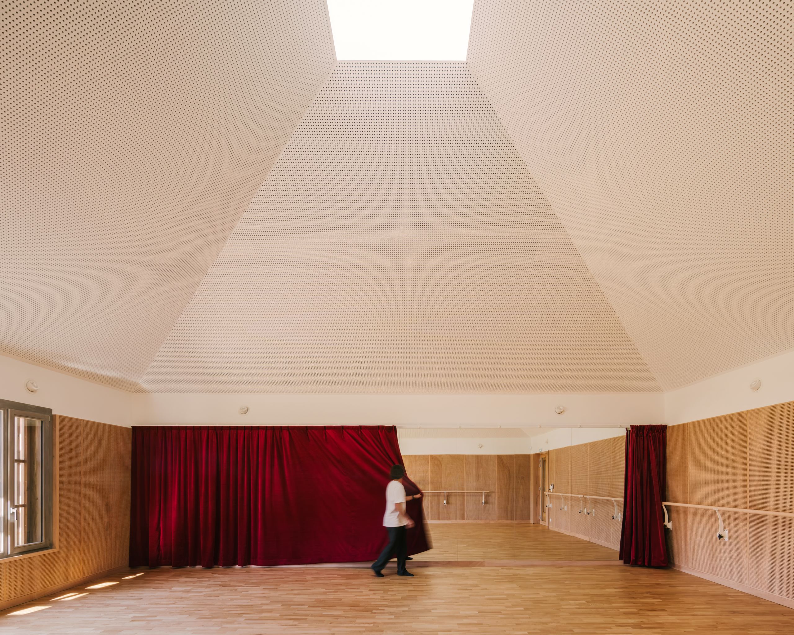 Dance studio of the Youth Cultural Centre in Vaison-la-Romaine, with timber floor, acoustic timber wall panels, red curtain and skylit faceted ceiling.