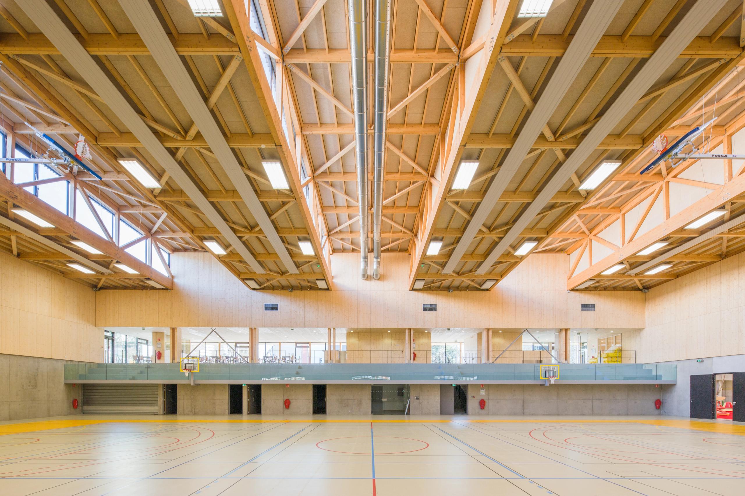 Intérieur du gymnase Hacine Cherifi à Rillieux-la-Pape, grande halle sportive en béton et bois, charpente treillis apparente et lumière naturelle zénithale.