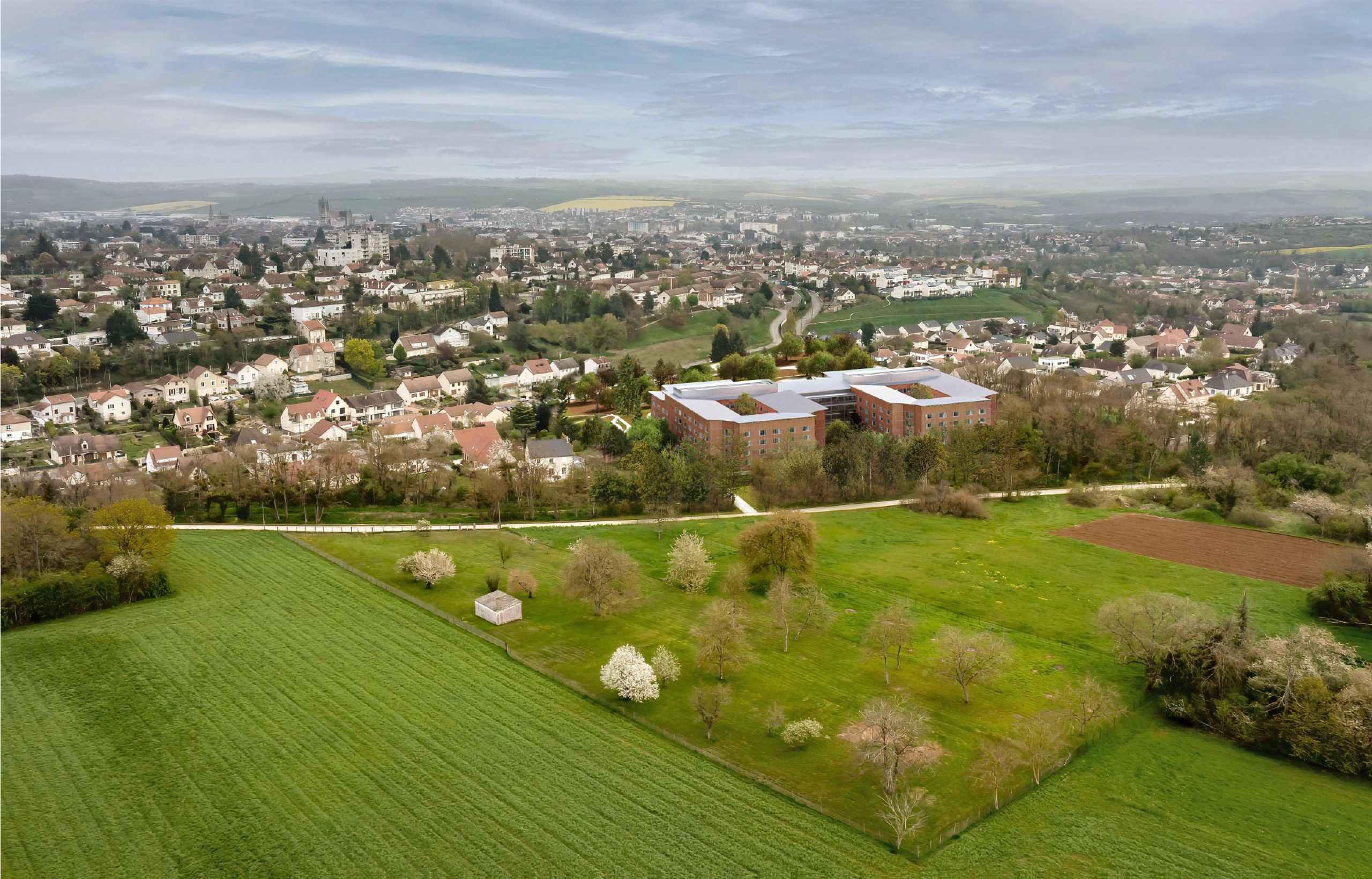 Vue aérienne de l’Ehpad d’Auxerre : deux pavillons en brique organisés autour de patios, implantés entre parc arboré, voie verte et tissu pavillonnaire.