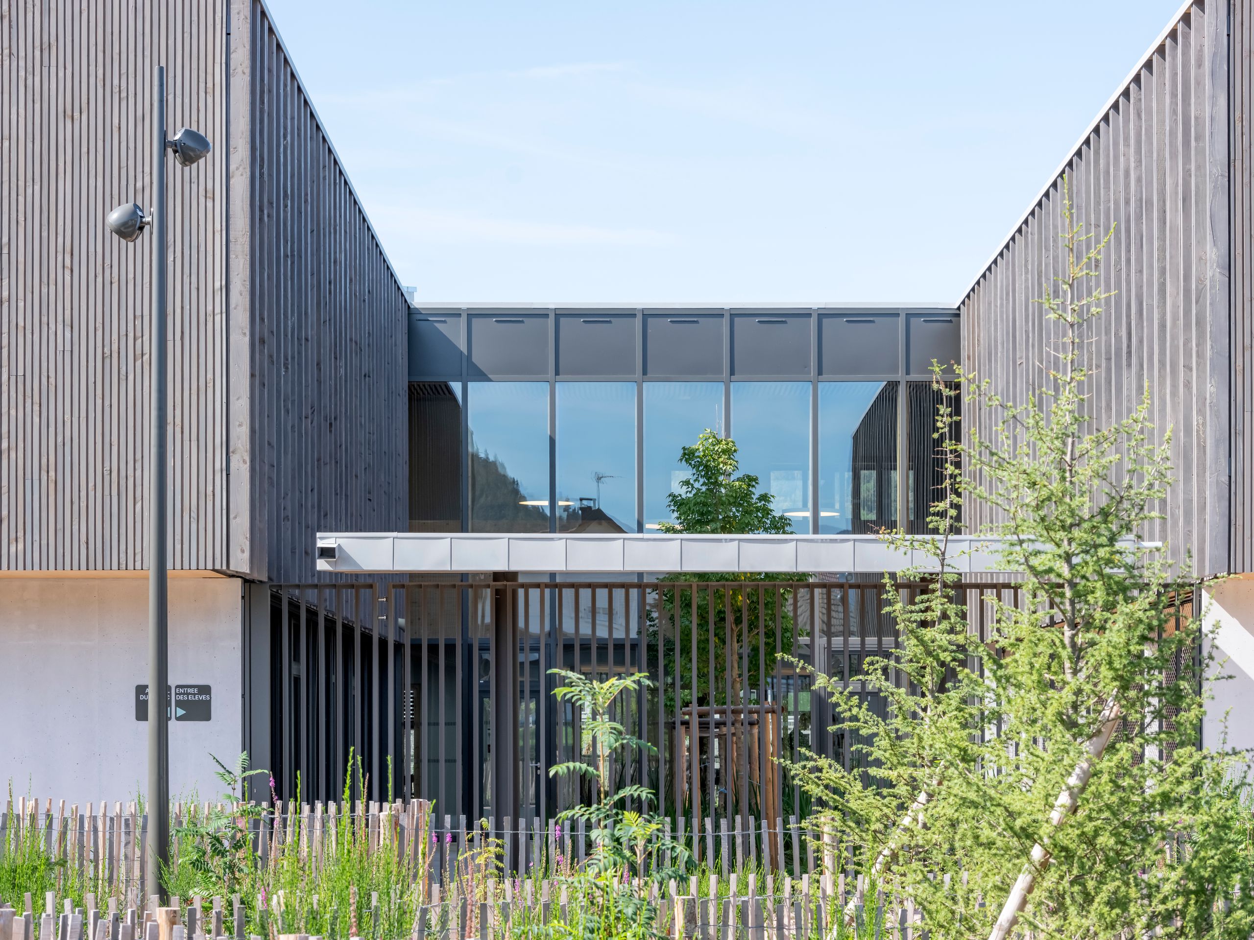 Façade du Collège du Ban à Vagney : volumes en bois vosgien, portique vitré sur cour plantée, structure en peigne et clôture paysagère au premier plan.