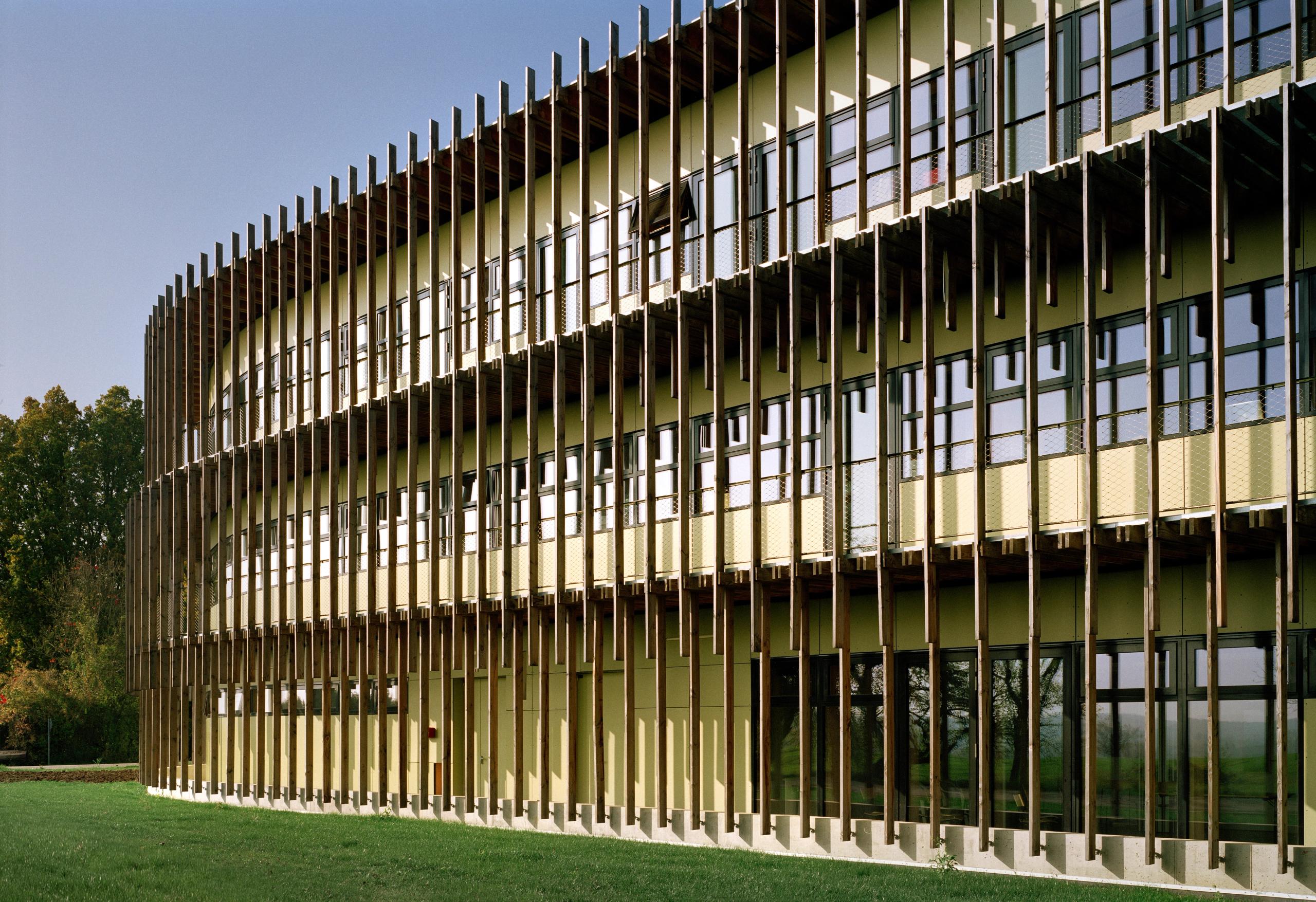 Façade sud courbe du laboratoire INRAE à Champenoux, bardage bois à lames verticales filtrant de larges baies vitrées sur trois niveaux, ouvrant sur le parc.