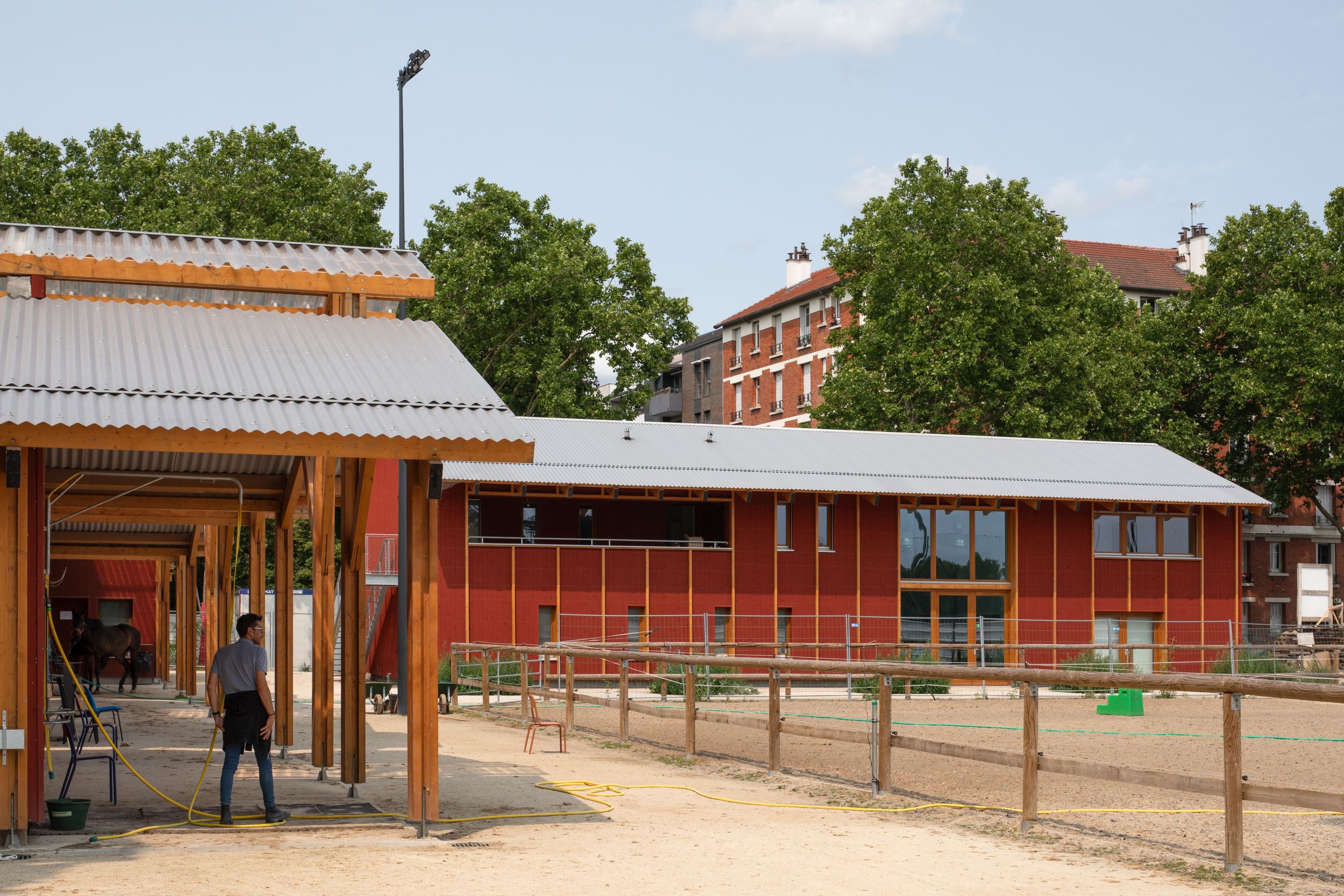 Centre équestre de Gennevilliers : bâtiments en ossature bois et bardage rouge de Falun, toitures en tôle ondulée encadrant les paddocks en milieu urbain dense.