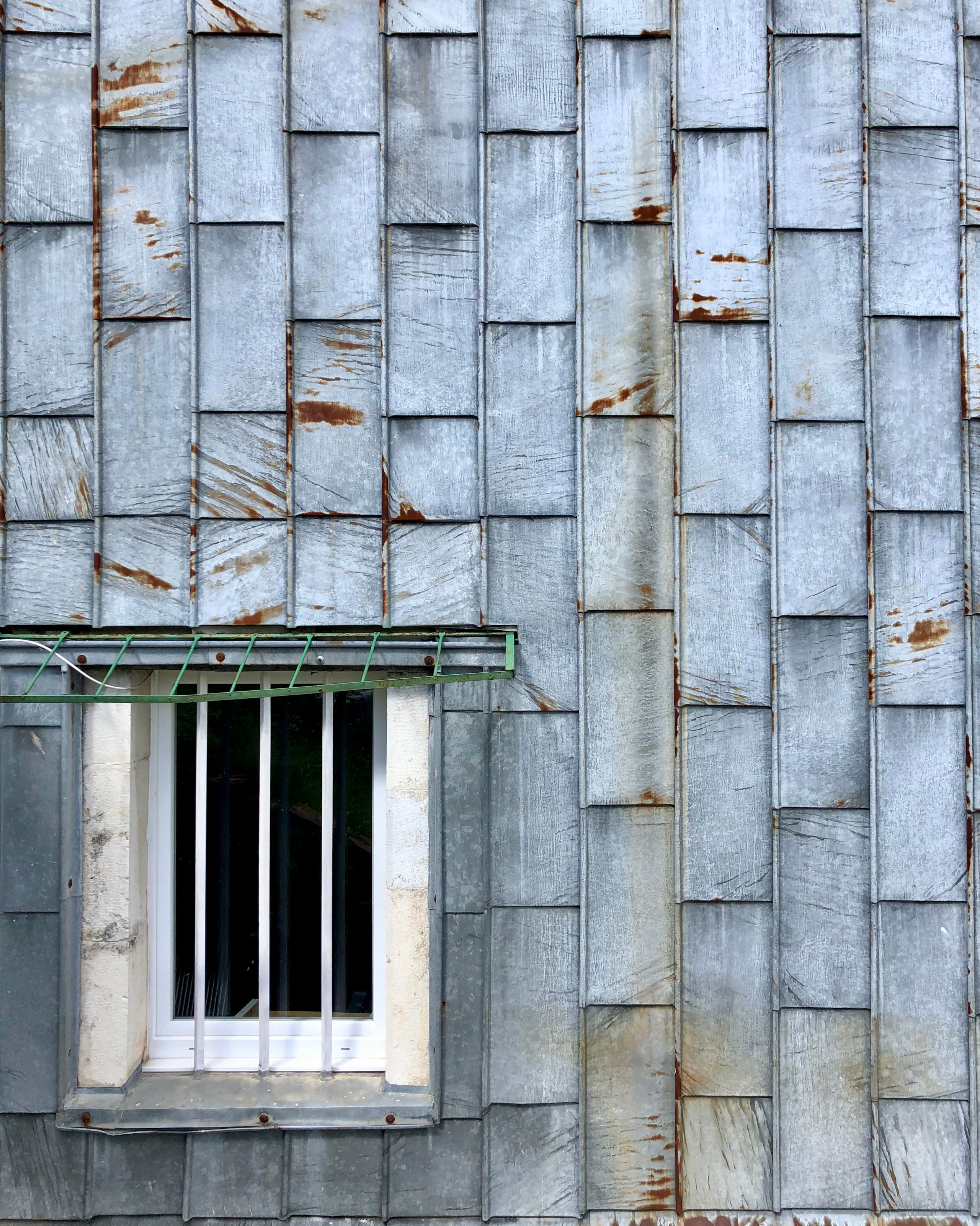 Façade en zinc patiné de l’école élémentaire Hubert Reeves à Champagnole, détail d’une fenêtre encadrée de pierre avec barreaux verticaux et auvent métallique.