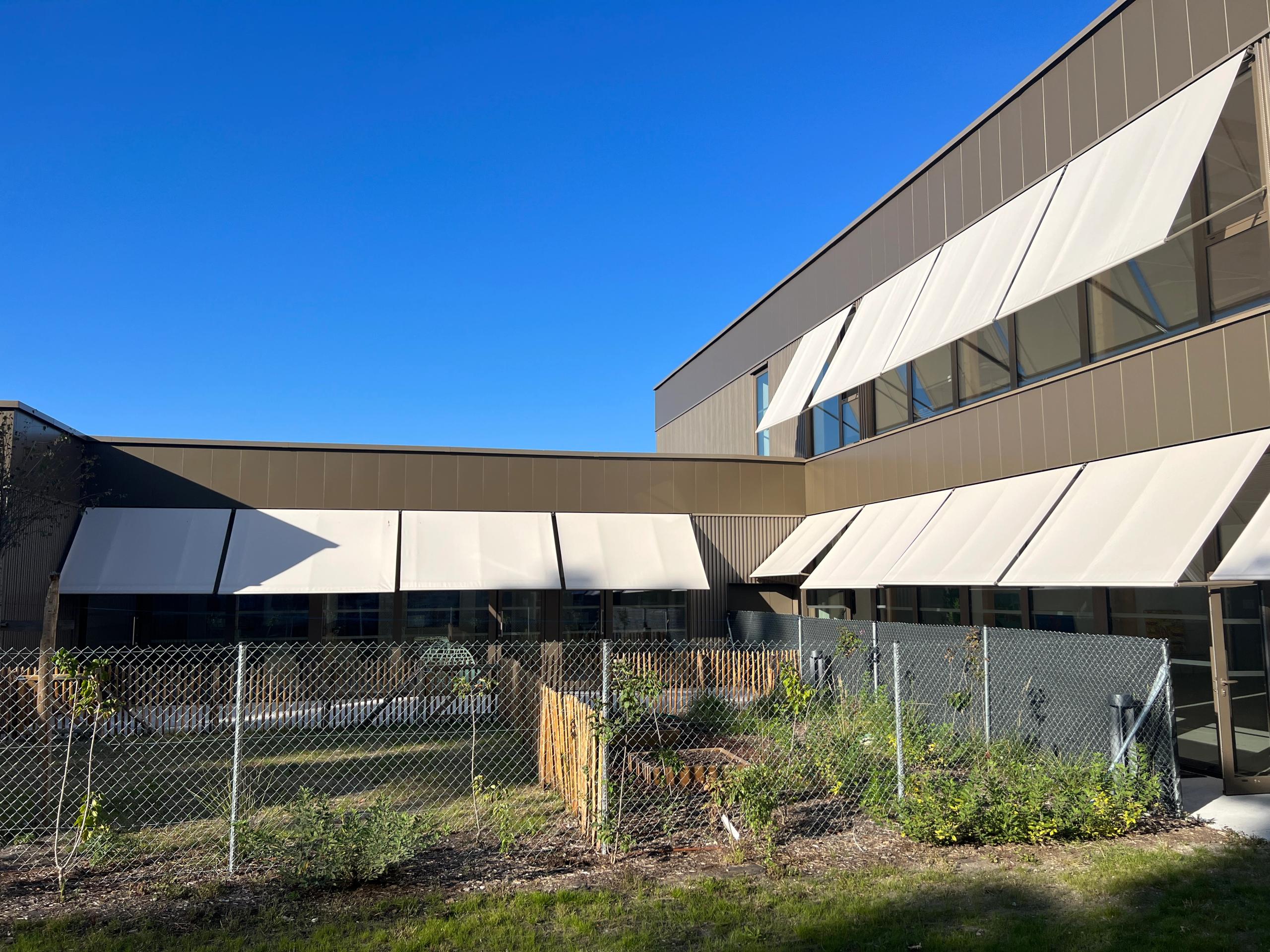 Vue de la maison des habitants restructurée et étendue à Mérignac par Tectoniques, façades métalliques beige, larges baies vitrées et stores extérieurs sur jardin clôturé.