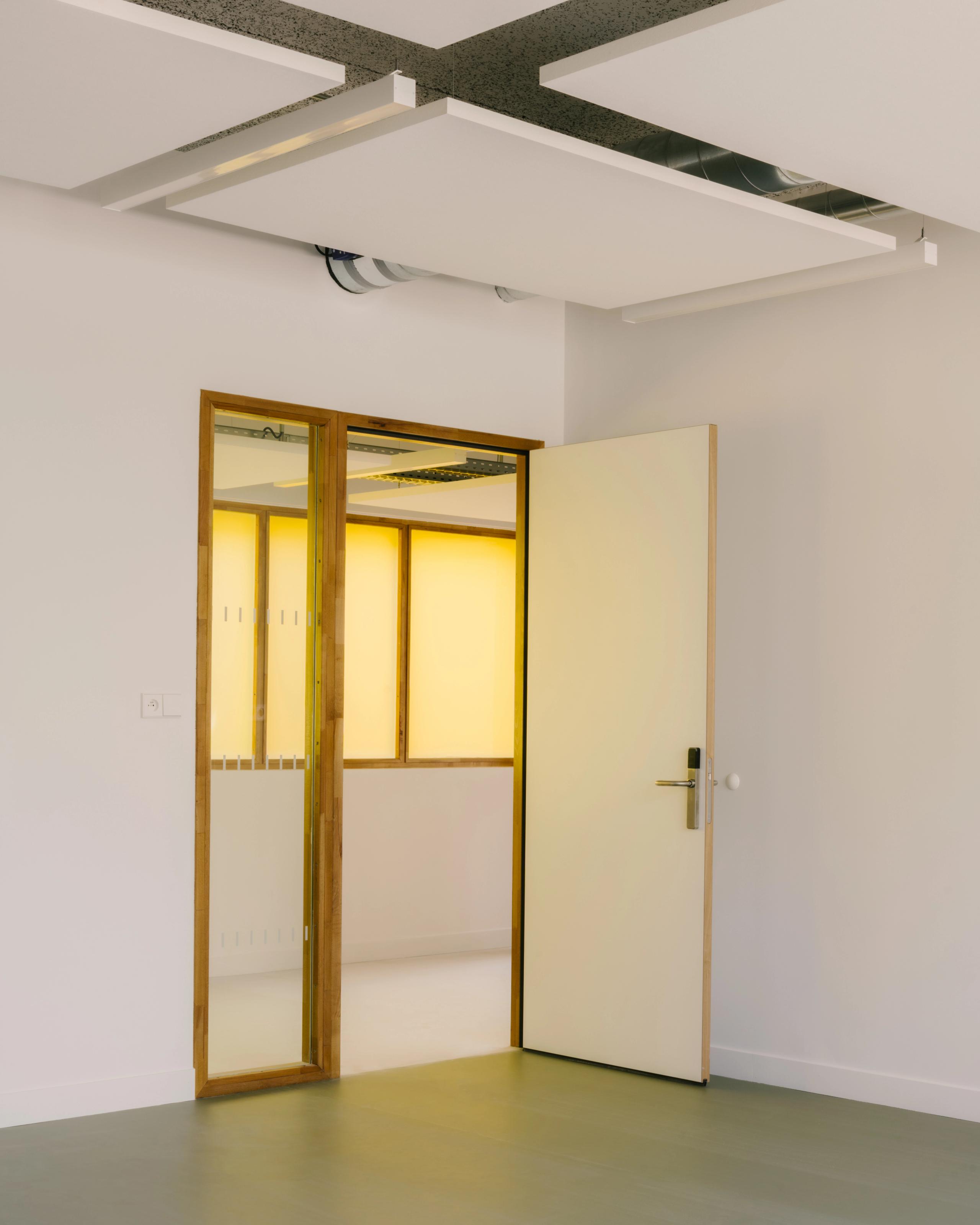 Intérieur de la Maison des familles à Limonest : porte bois-verre ouvrant sur un couloir baigné de lumière jaune, plafond technique avec dalles suspendues.