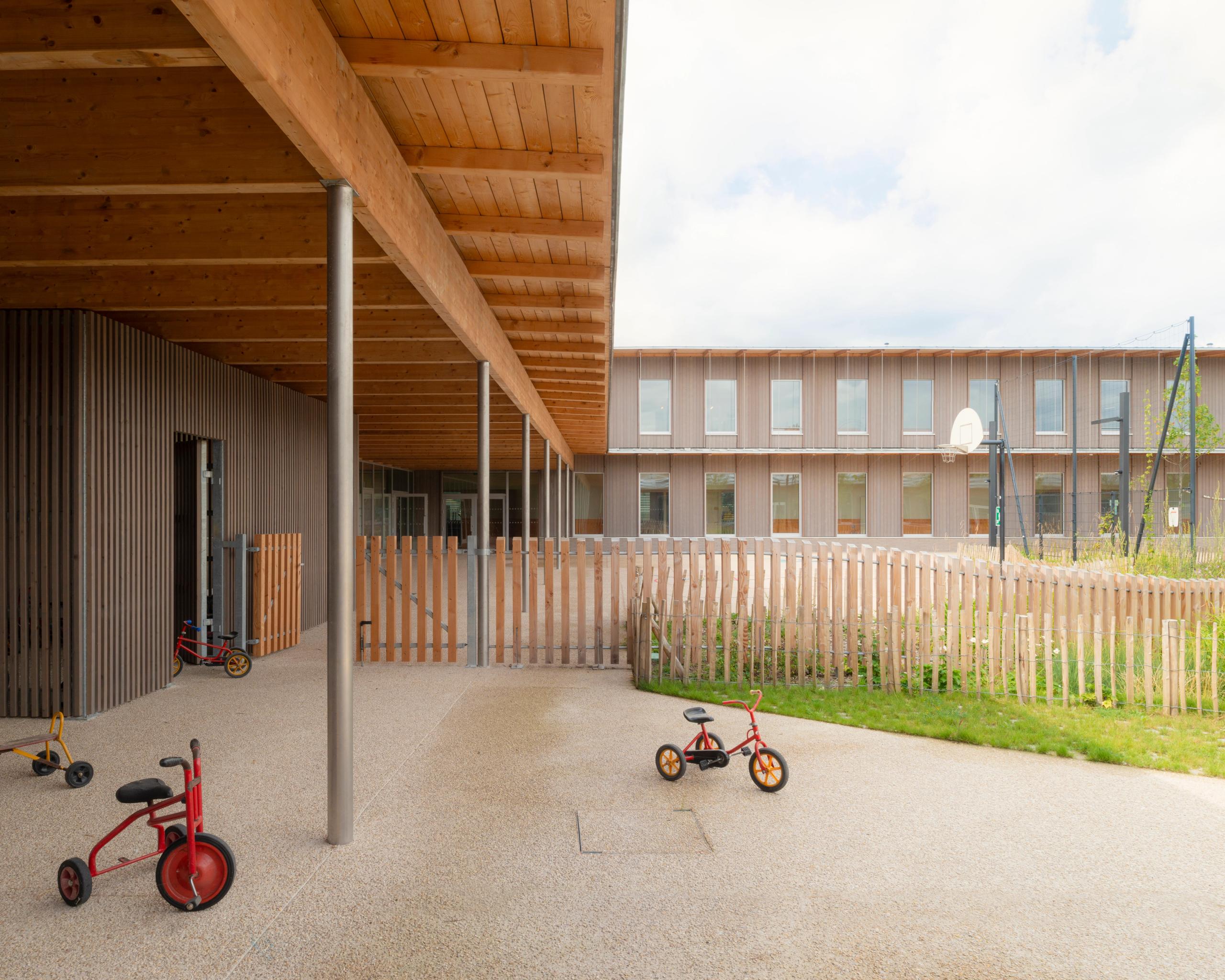 Cour des groupes scolaires Jacqueline Auriol et Jean Jaurès au Bourget, préau en ossature bois, poteaux métalliques et façades bois rythmées sur deux niveaux.