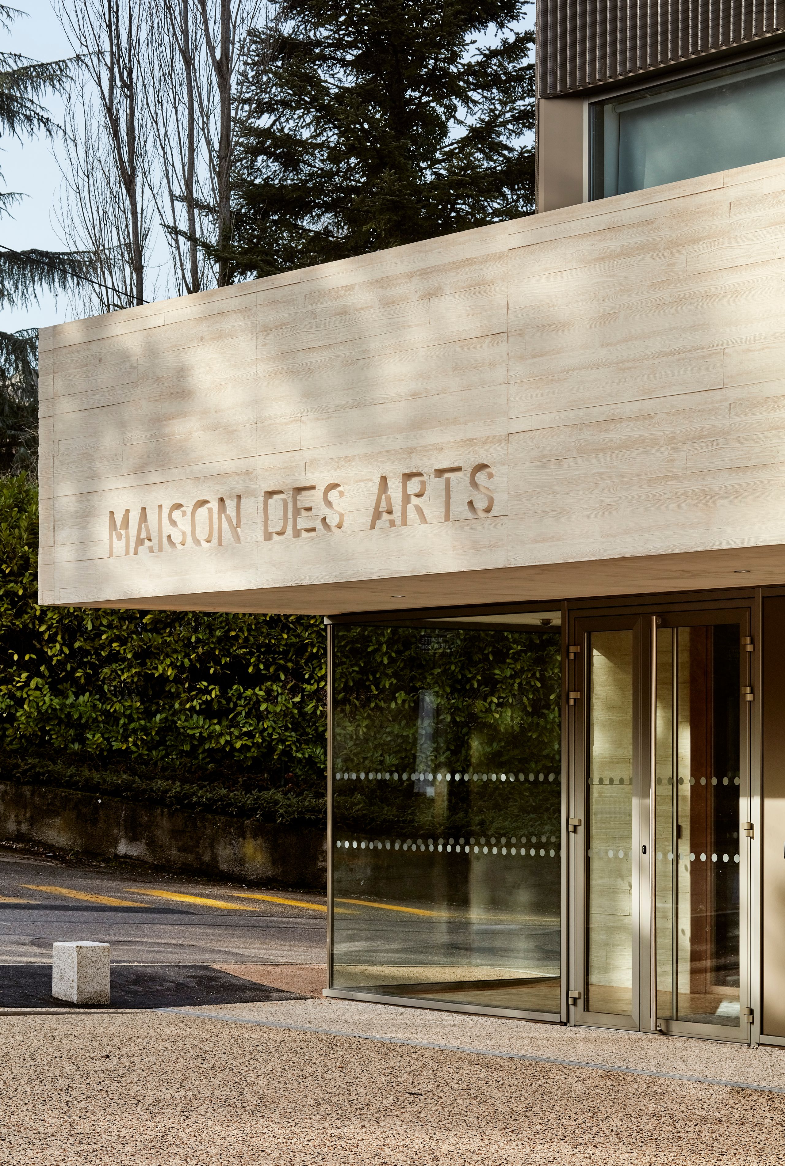 Entrée de la Maison des Arts à Charbonnières-les-Bains, volume en béton clair en porte-à-faux sur façade vitrée, en lien avec le paysage arboré en arrière-plan.