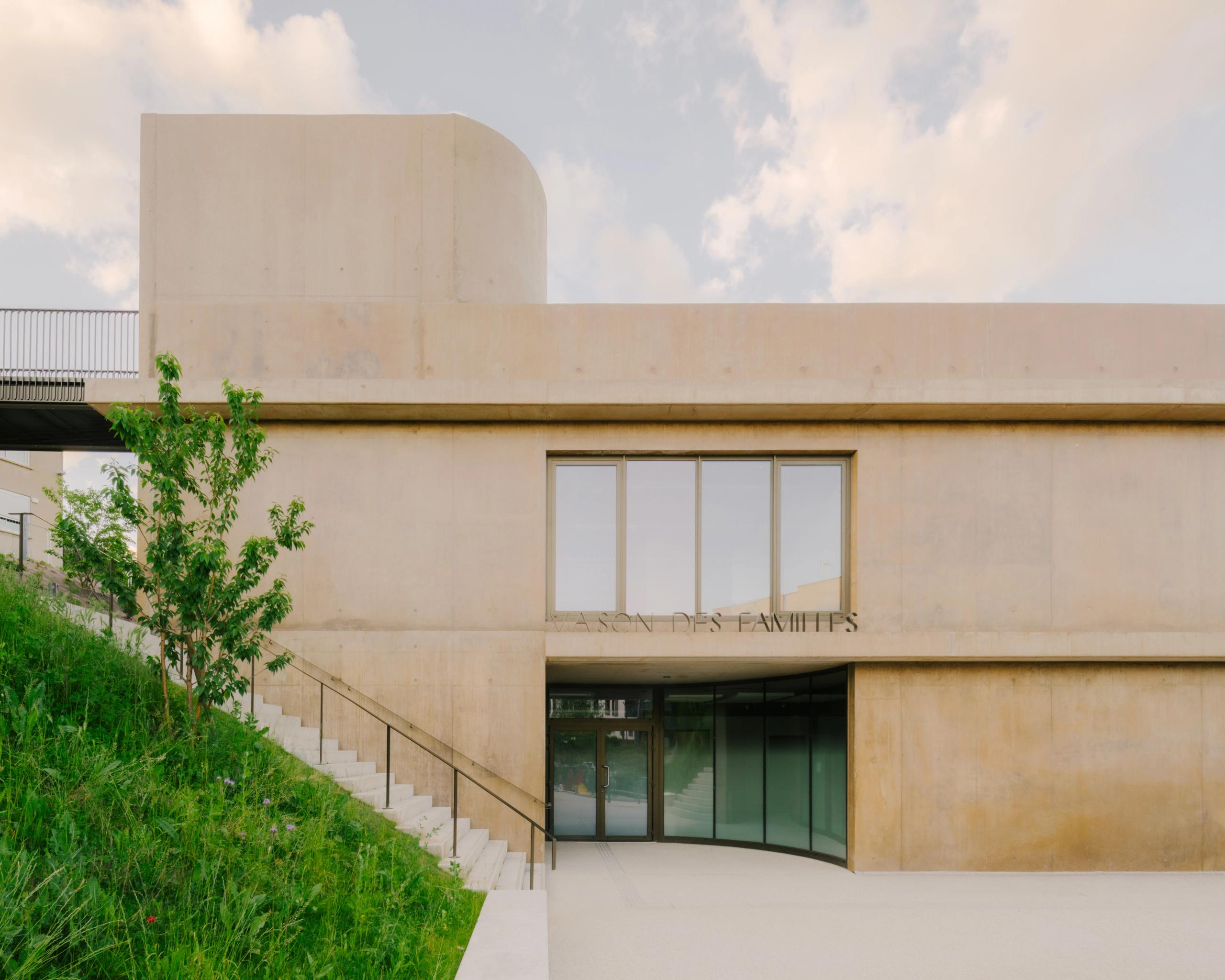 Façade en béton teinté de la Maison des familles à Limonest, volume en strates avec entrée vitrée en retrait et escalier extérieur longeant le talus végétalisé.
