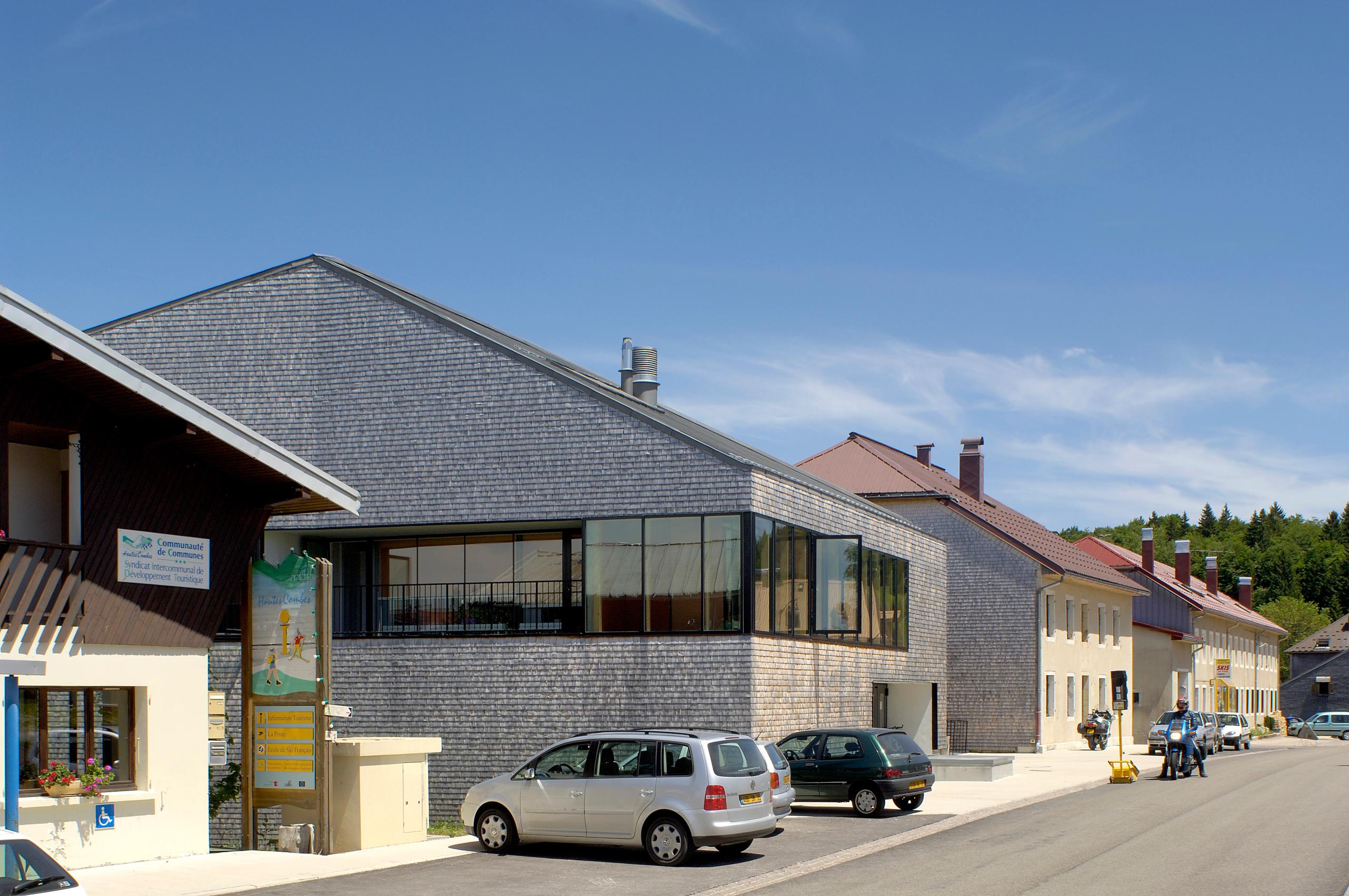 Maison du Parc du Haut-Jura à Lajoux, volume compact en tavaillons d’épicéa et bande vitrée continue sur rue, inséré dans le front bâti existant.