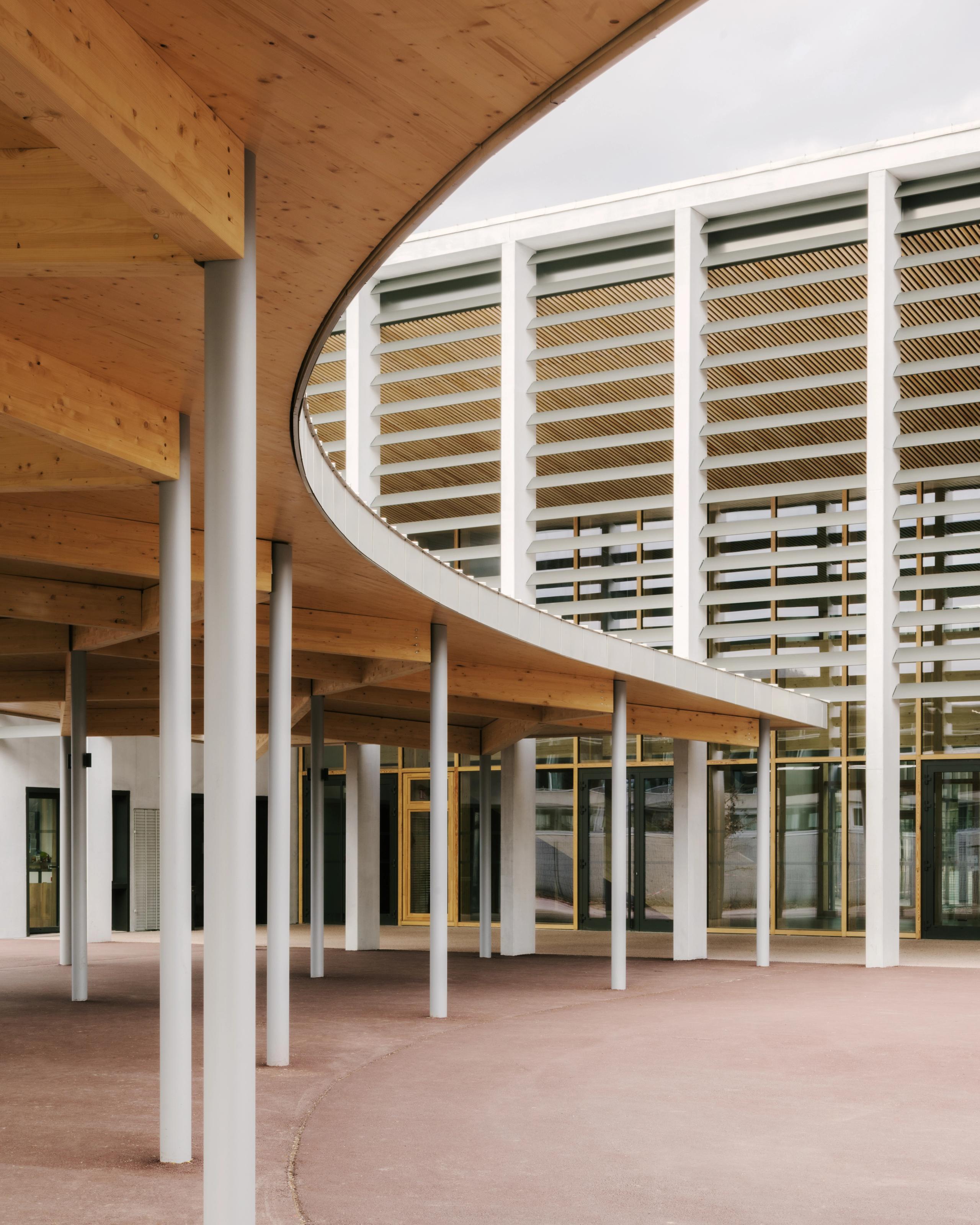 Préau courbe en bois et poteaux fins du Collège du Ban à Vagney, ouvrant sur une façade rythmée de brise-soleil et menuiseries bois-métal.