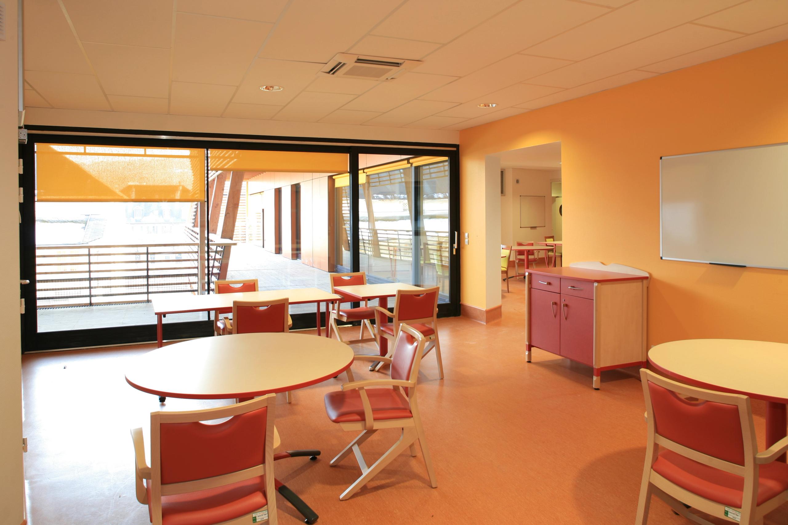 Espace de vie de l’EHPAD de Chambéry, salle commune ouverte sur une large terrasse bois, menuiseries vitrées coulissantes et protections solaires extérieures.
