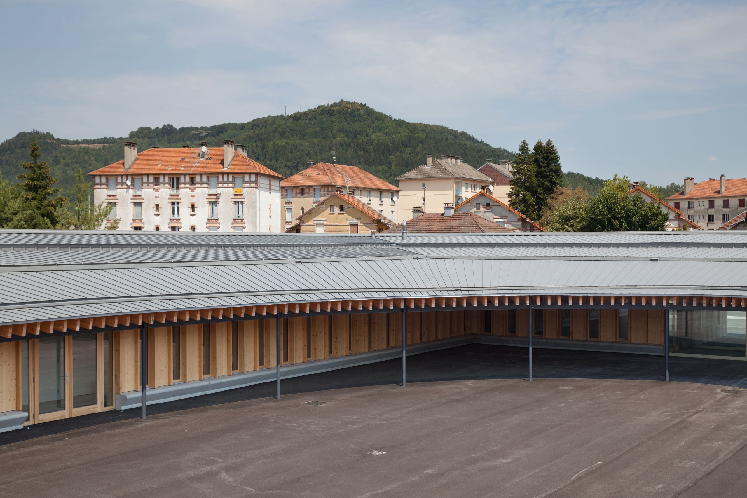 Cour elliptique de l’école élémentaire Hubert Reeves à Champagnole, entourée d’un volume bas en ossature bois et toiture zinc, sur fond de tissu urbain et relief boisé.
