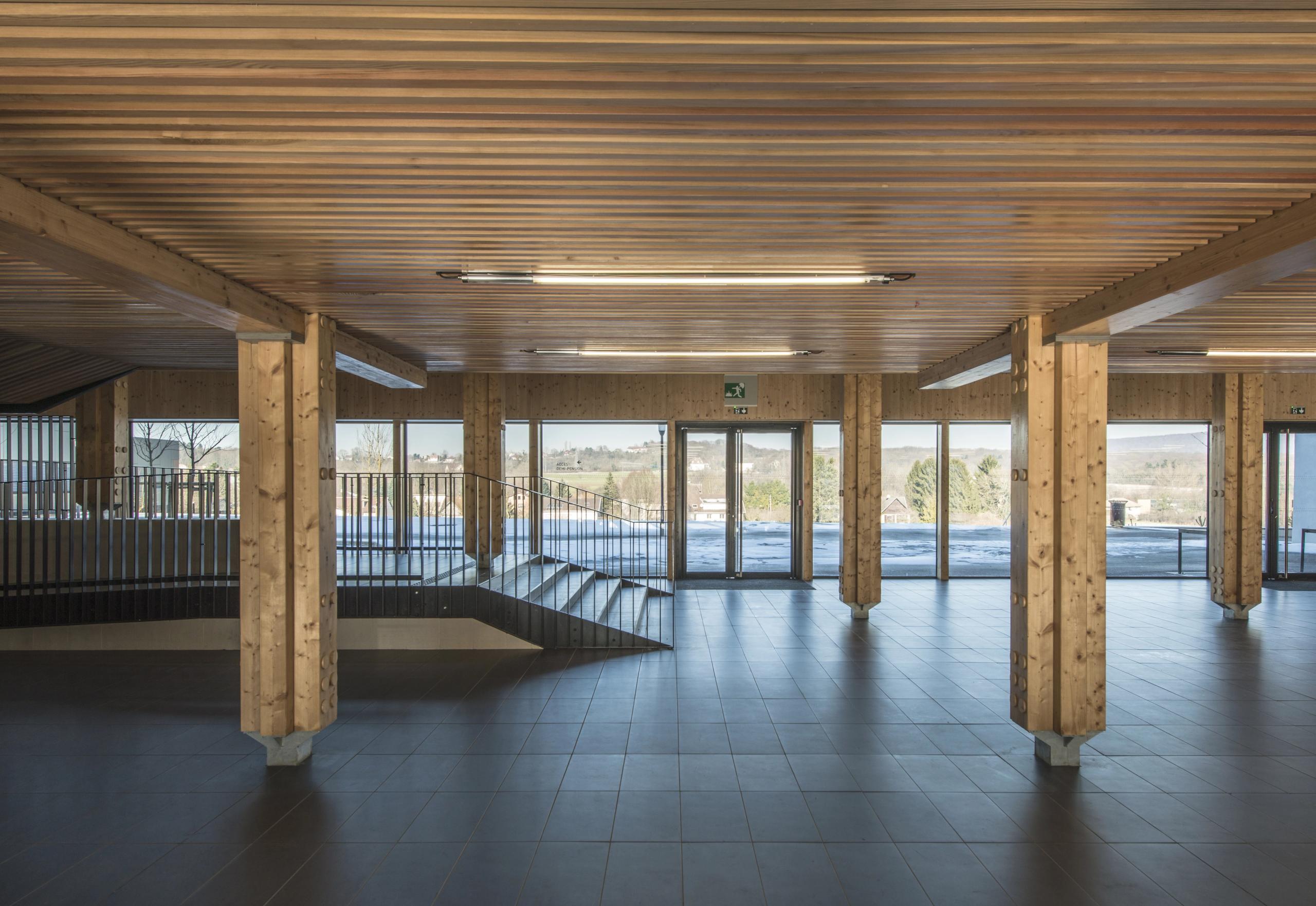 Hall traversant du collège Pierre-Joseph Proudhon à Besançon, structure poteau-poutre bois apparente, sol carrelé sombre et larges baies vitrées sur le paysage.