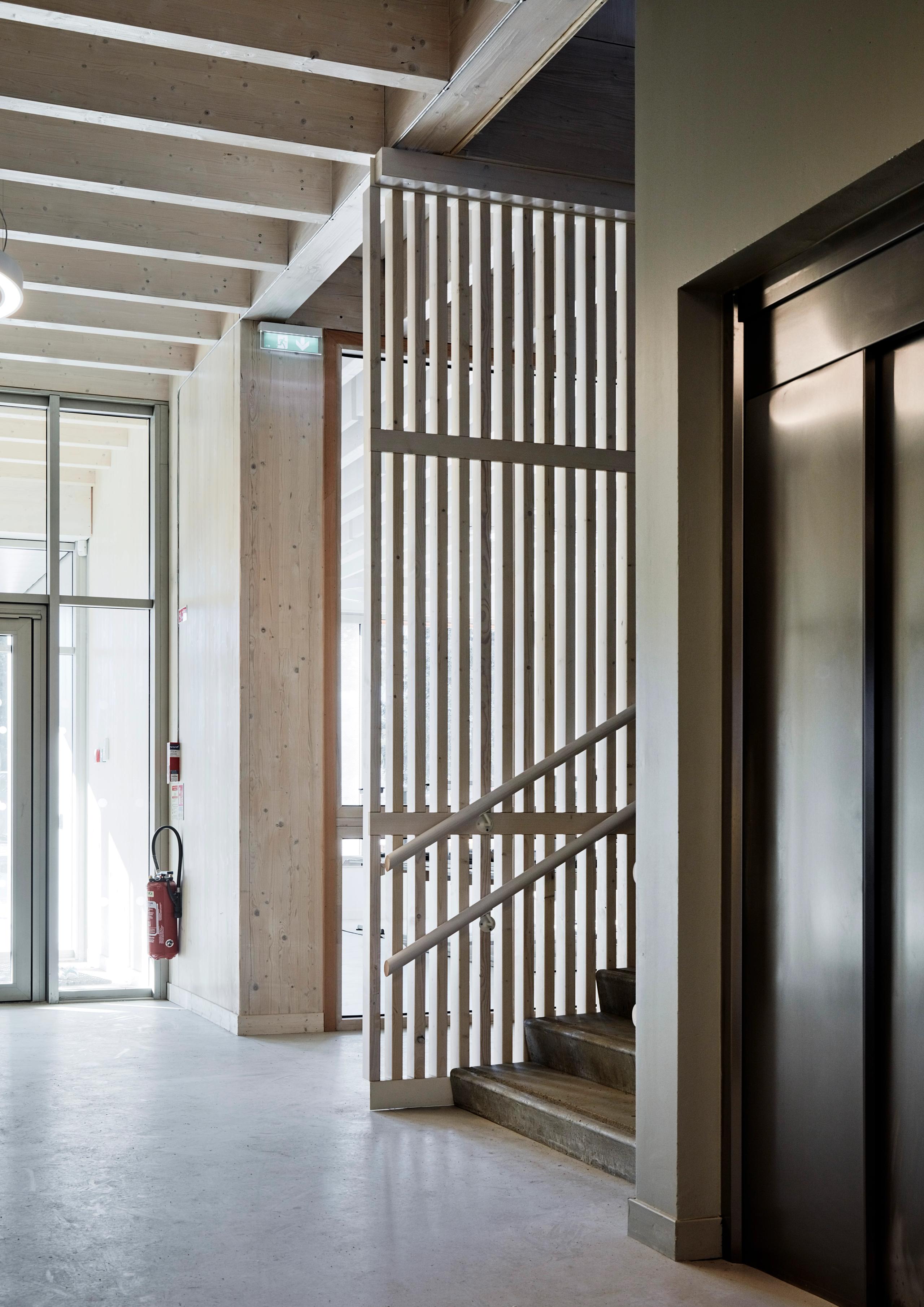 Hall de la Maison de l’Enfance Simone Veil à Albertville, avec structure bois apparente, escalier béton filtré par claustra et ascenseur métallique.