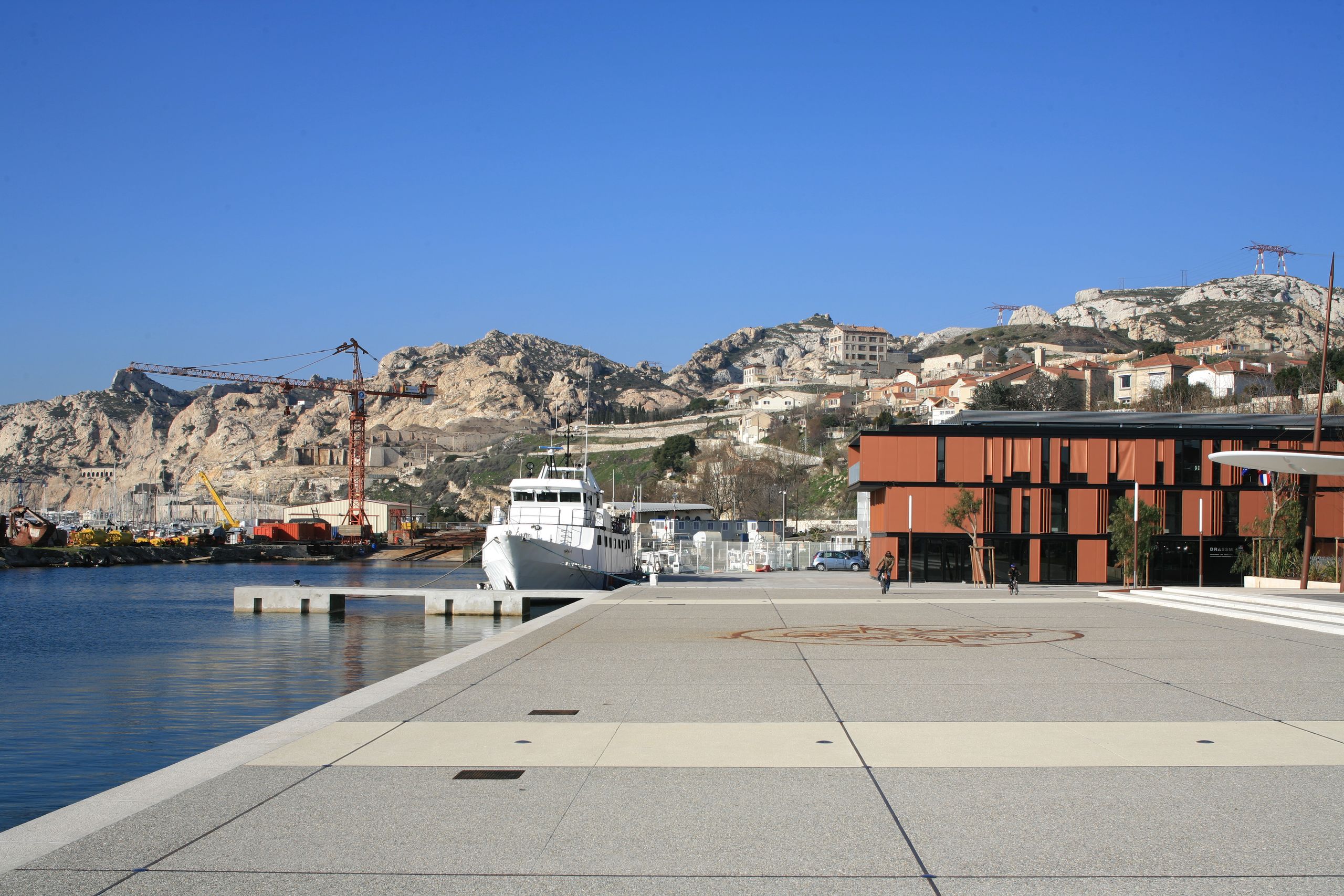 Façade en panneaux métalliques rouille du Département des Recherches Subaquatiques et Sous-Marines à Marseille, ouvrant sur un vaste parvis minéral en bord de quai.