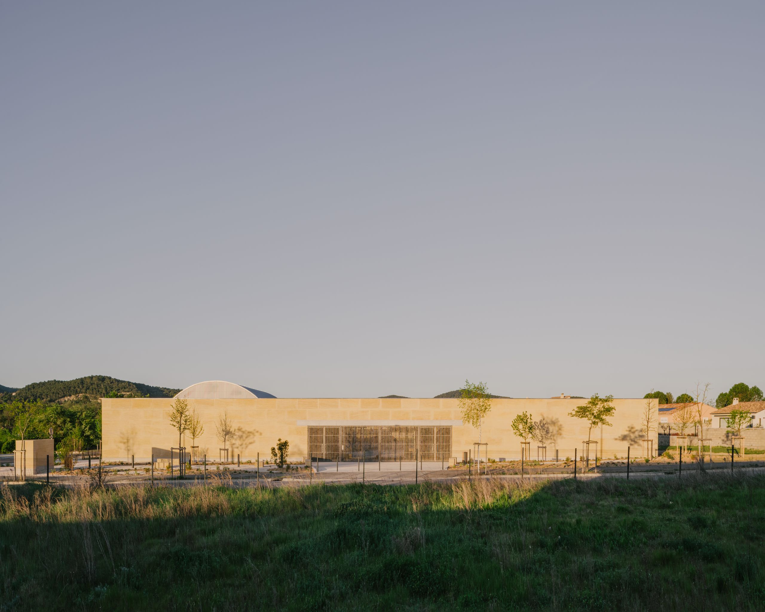 Street-side view of the Youth Cultural Centre in Vaison-la-Romaine, a low stone volume with metal screens and young trees set against distant hills.