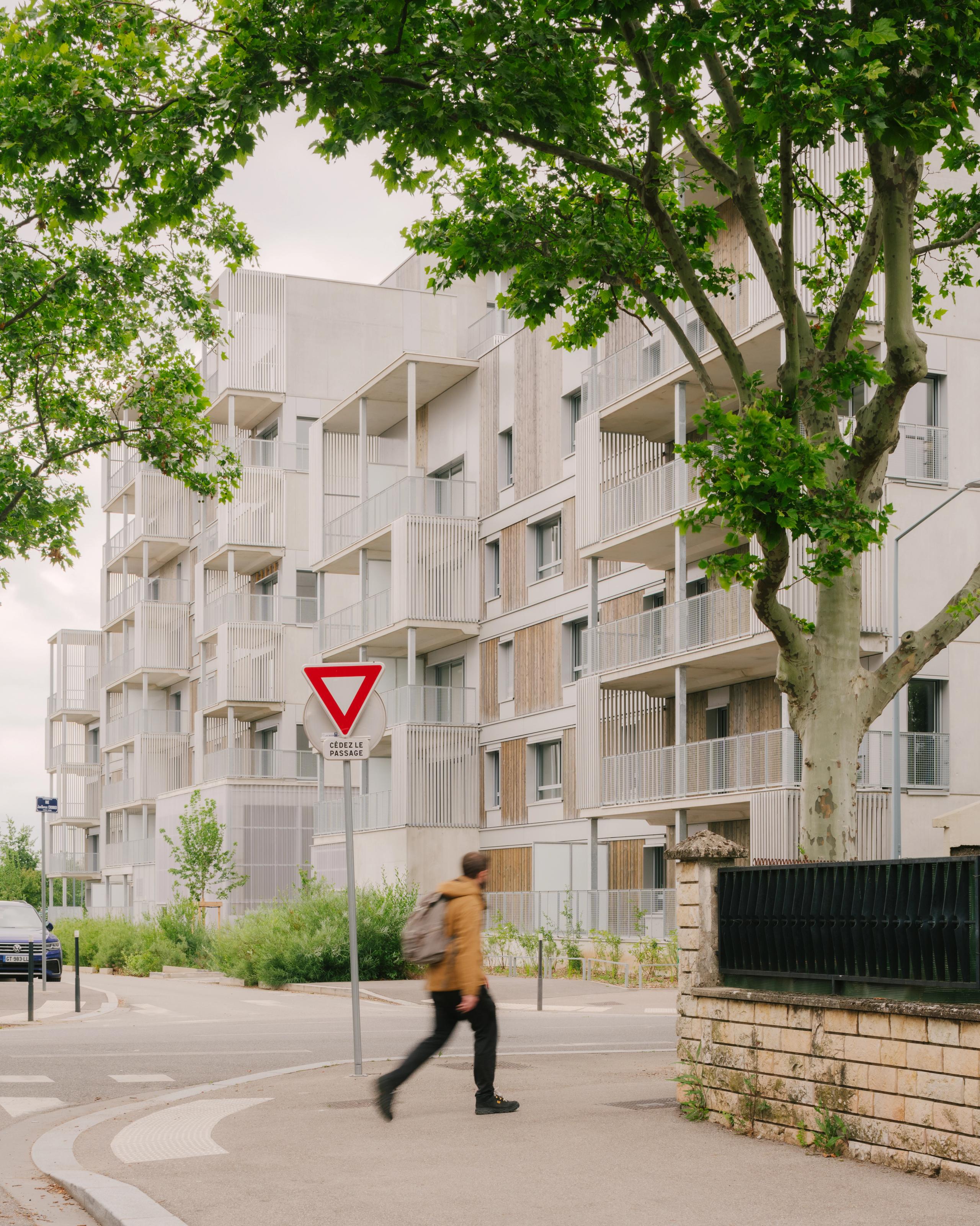 Vue de la résidence de 116 logements à Vénissieux, façades bois-béton fragmentées, balcons en exosquelette métallique et socle paysager en pied d’immeuble.