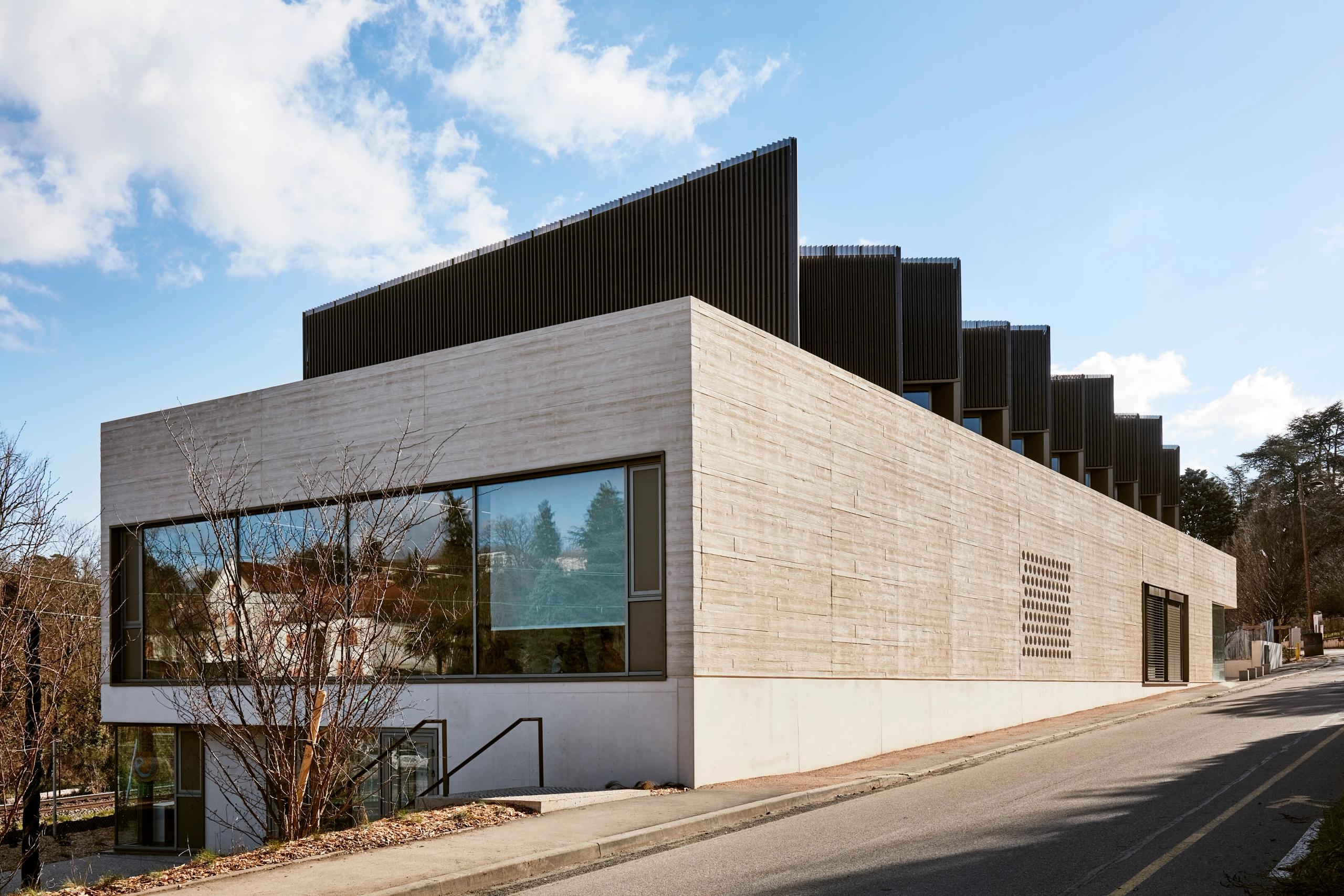 Maison des Arts à Charbonnières-les-Bains : volume en béton matricé en socle, couronnement en bois et métal perforé, façade vitrée sur rue en pente.