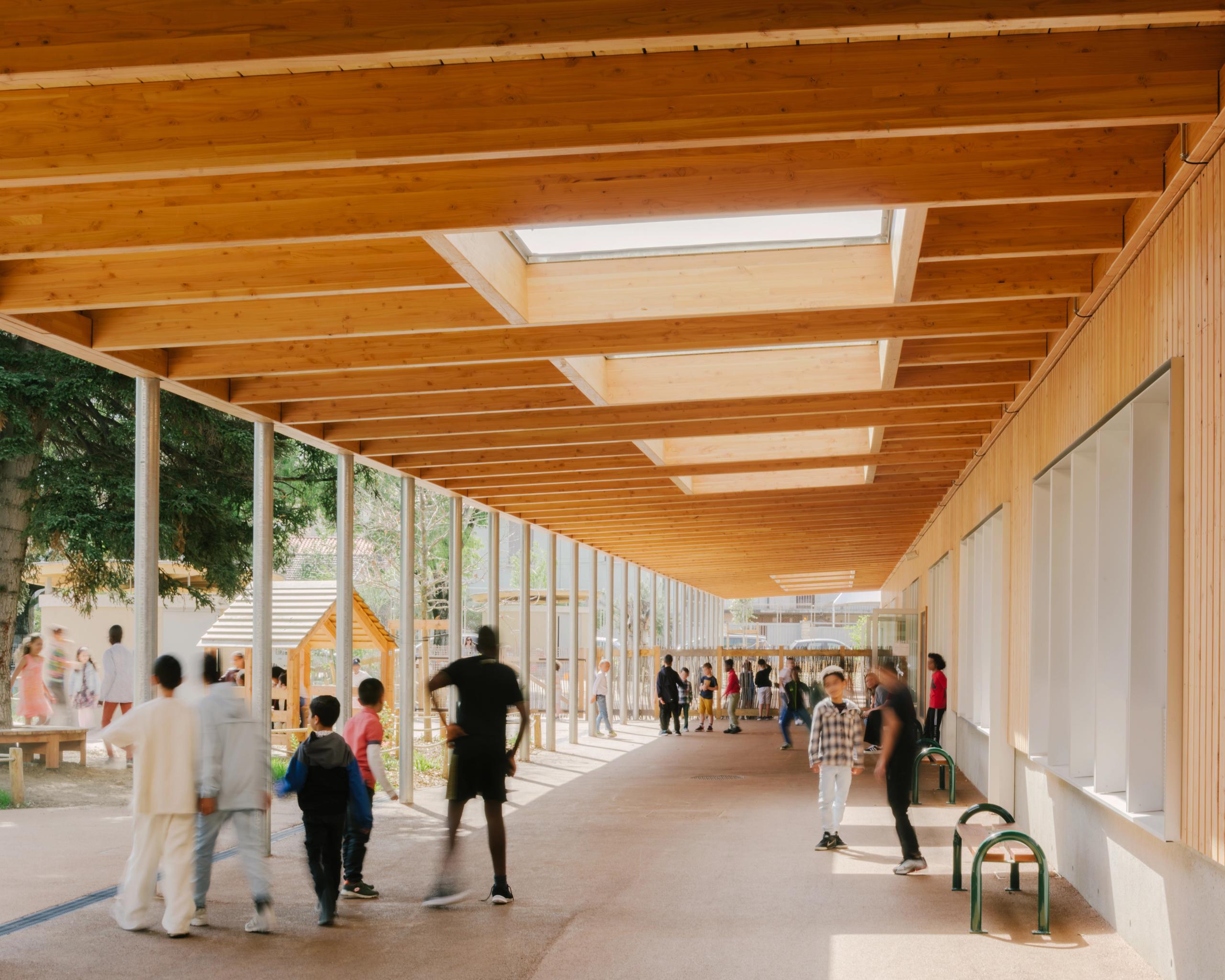 Galerie couverte en bois du groupe scolaire Kennedy à Lyon, structure poteaux-poutres ouverte sur la cour végétalisée, éclairée par de grands lanterneaux.