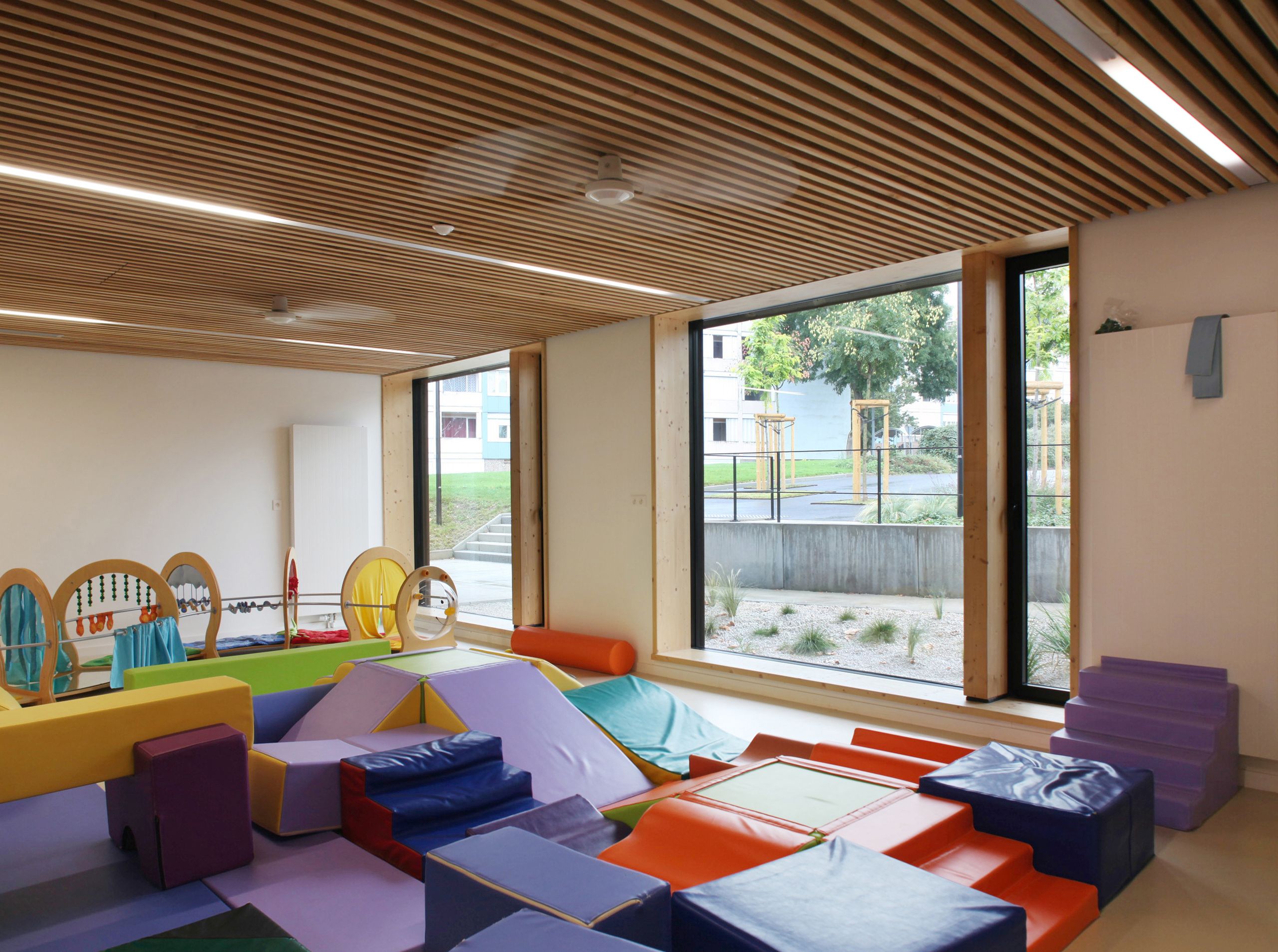 Espace de motricité de la Maison de la petite enfance à Autun, avec plafond à lames bois, grandes baies vitrées sur le jardin et modules souples colorés.