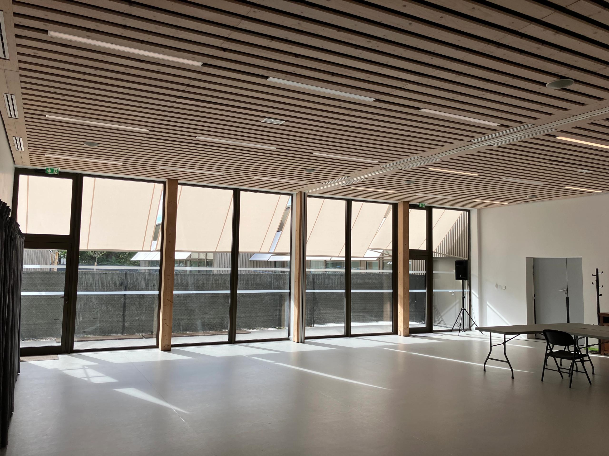 Salle polyvalente de la maison des habitants à Mérignac, livrée, avec façade vitrée toute hauteur, brise-soleil extérieurs et plafond bois à lames ajourées.