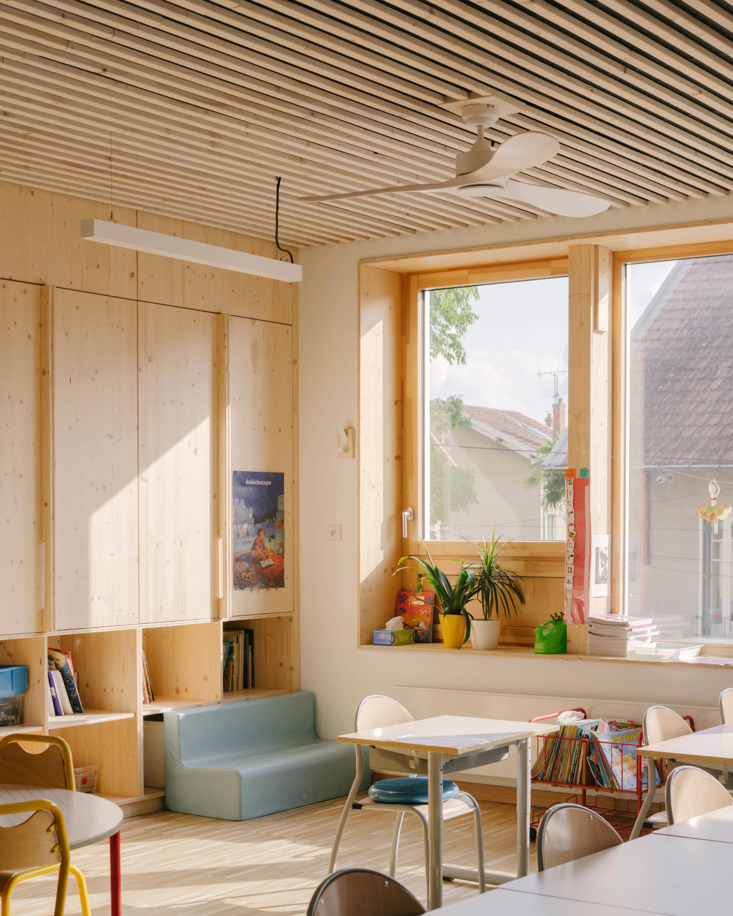 Salle de classe du Groupe scolaire Kennedy à Lyon, structure bois apparente, plafond à lames, grandes baies vitrées et mobilier enfant adapté.