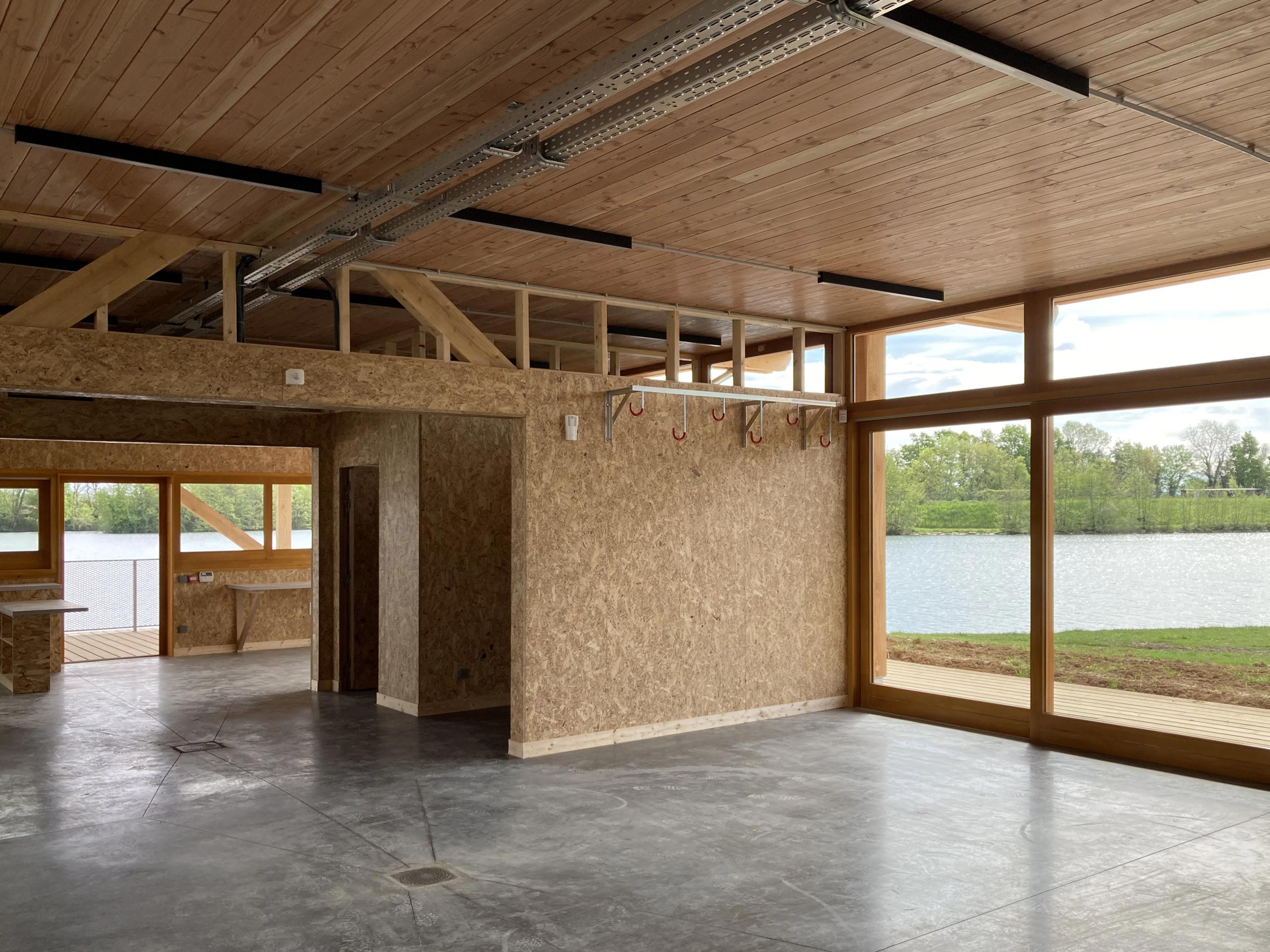 Intérieur de la Maison des sports à Malafretaz : structure bois apparente, parois en OSB, dalle béton et larges baies vitrées ouvrant sur le plan d’eau.