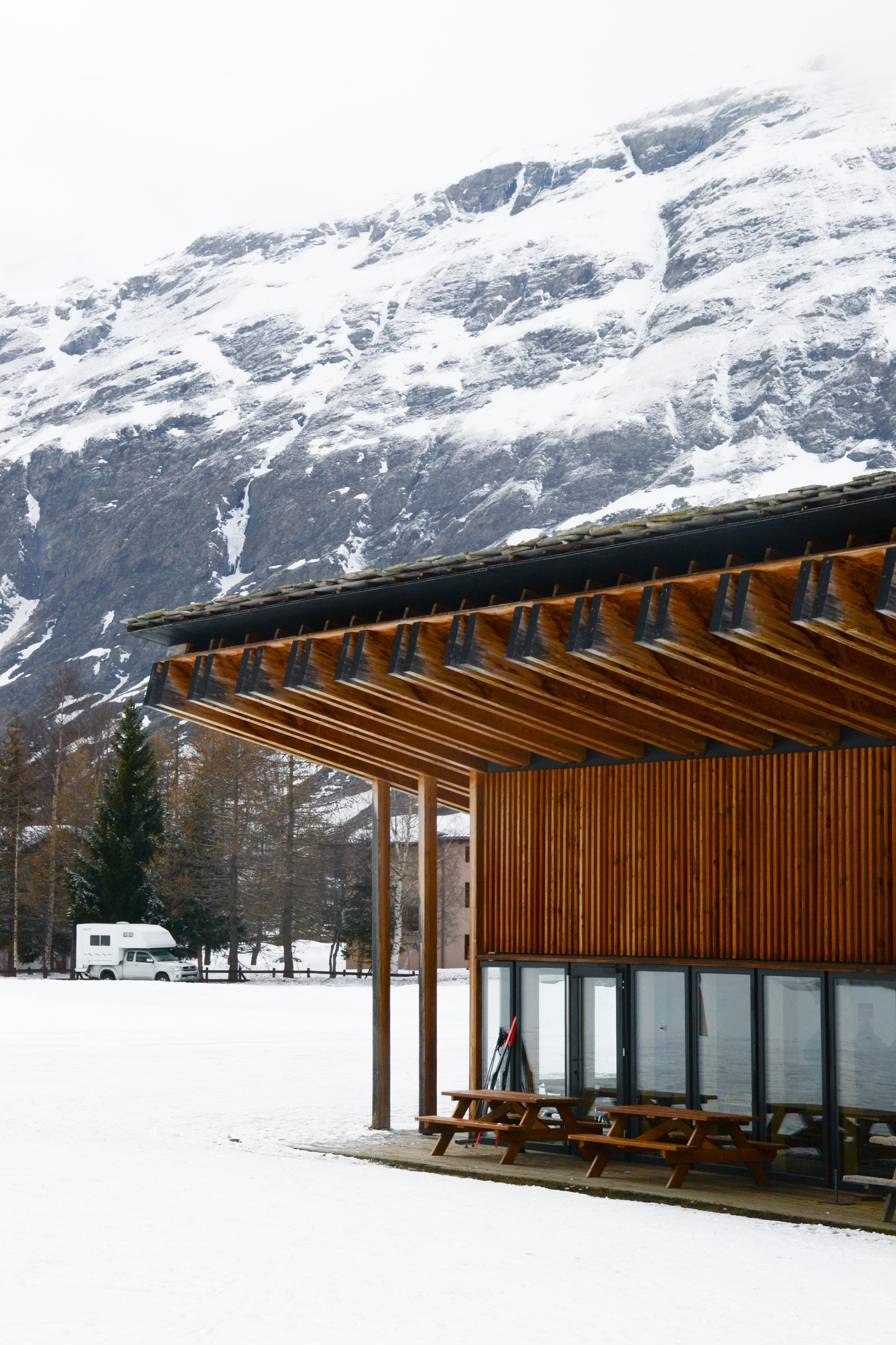 Espace nordique de Bessans : volume en bois à claire-voie, large auvent structurel et toiture en lauze, ouvert sur le paysage enneigé et le massif alpin.