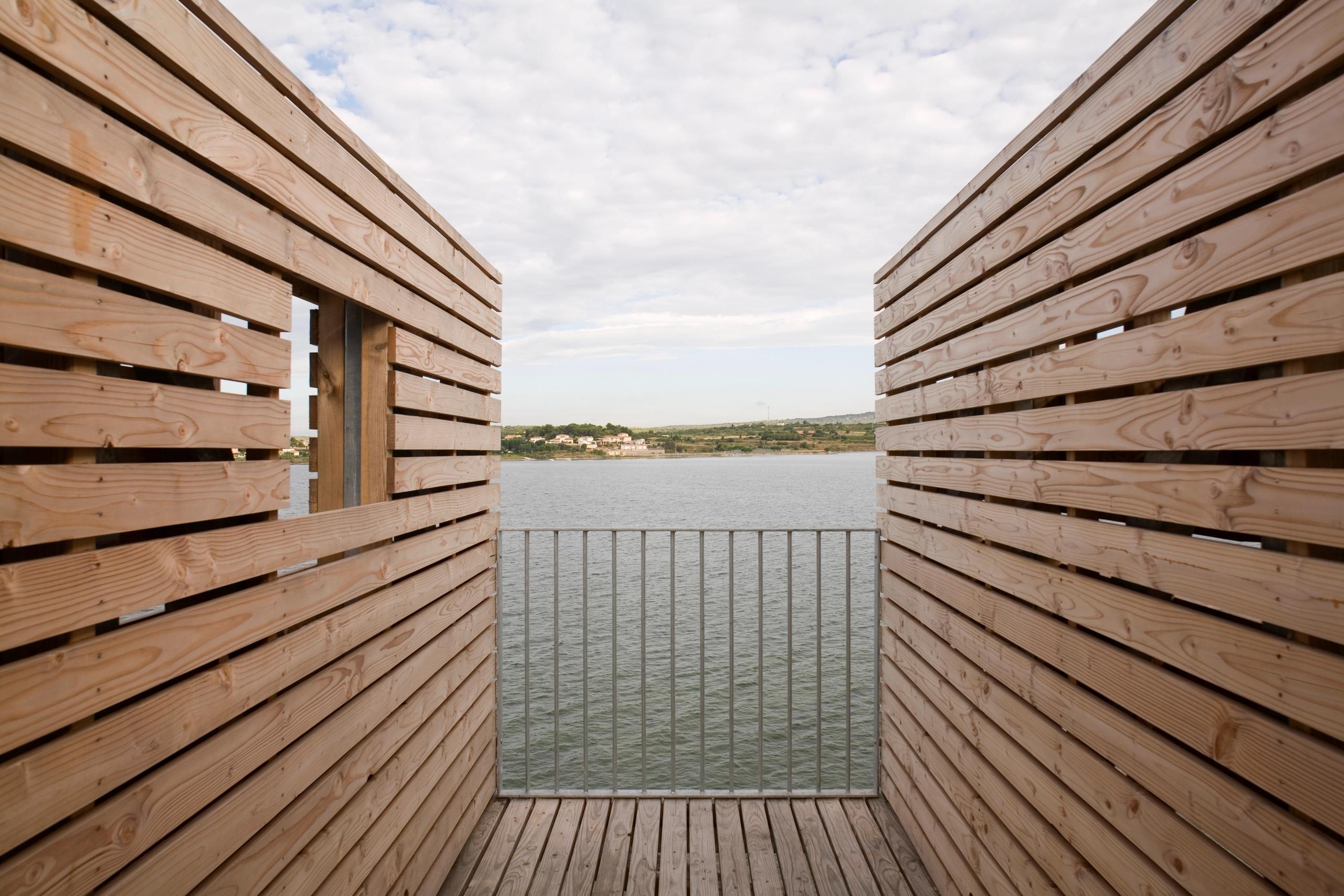 Passerelle en bois du centre thermal de Balaruc-les-Bains, encadrée de parois à claire-voie ouvrant une vue frontale sur l’étang et le paysage distant.