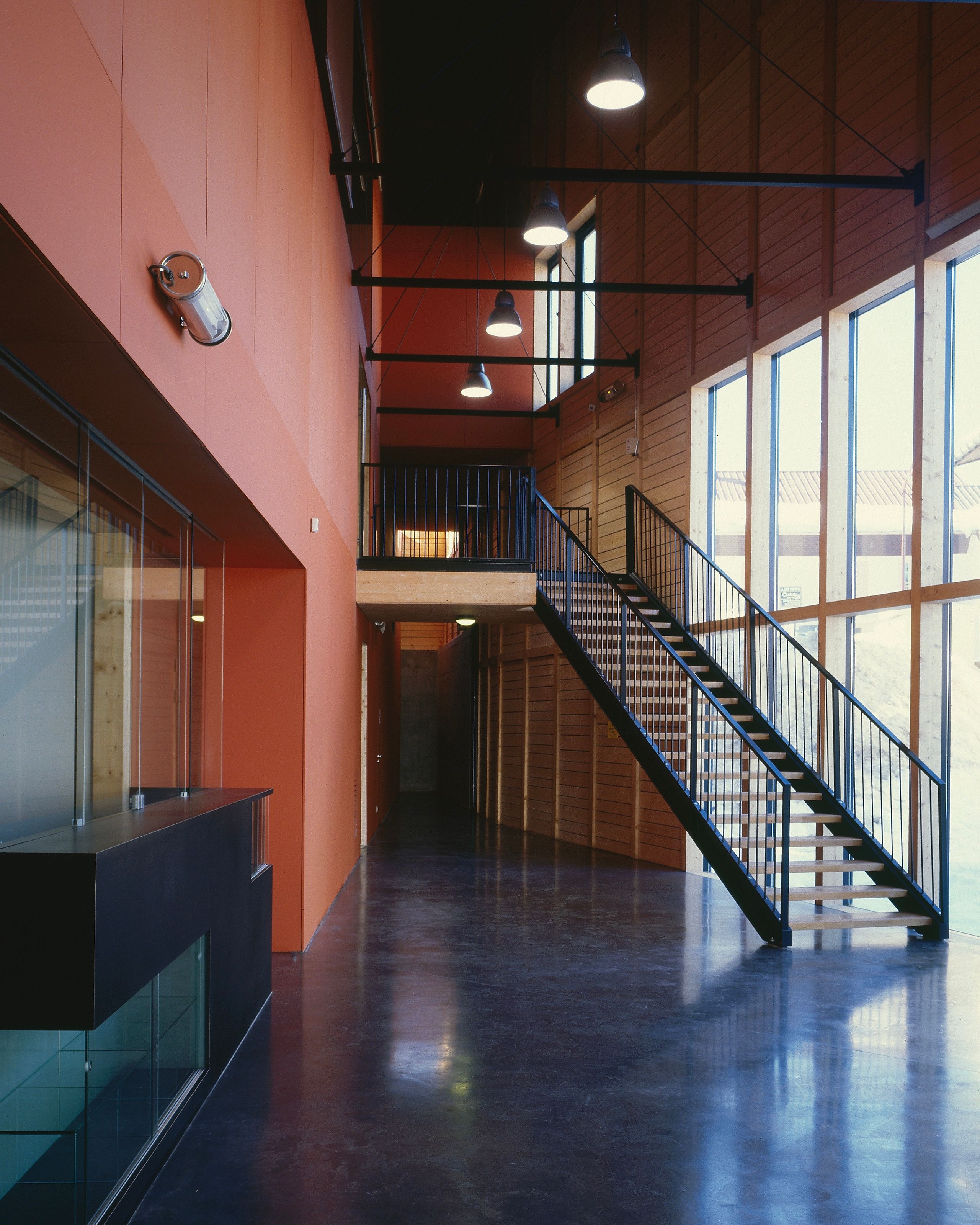 Hall de la Maison du Parc du Haut-Jura à Lajoux, double hauteur en bois, grande façade vitrée et escalier métallique noir sur sol poli sombre.