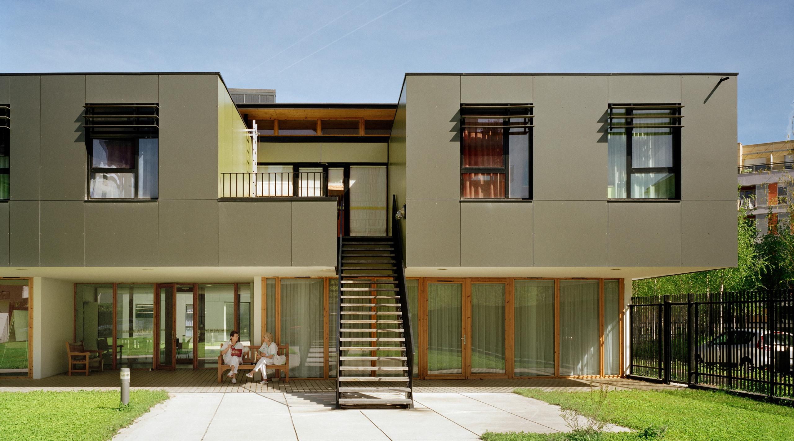 Façade de l’EHPAD à Grenoble par Tectoniques, volume R+1 en porte-à-faux, brise-soleil horizontaux, menuiseries bois vitrées sur jardin accessible.