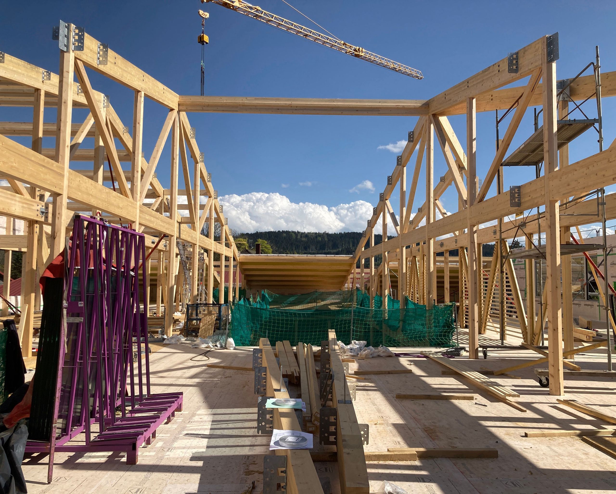 Chantier Tectoniques à [ville] : structure bois apparente en portiques et contreventements, plancher massif, grue et échafaudages sur fond de paysage boisé.