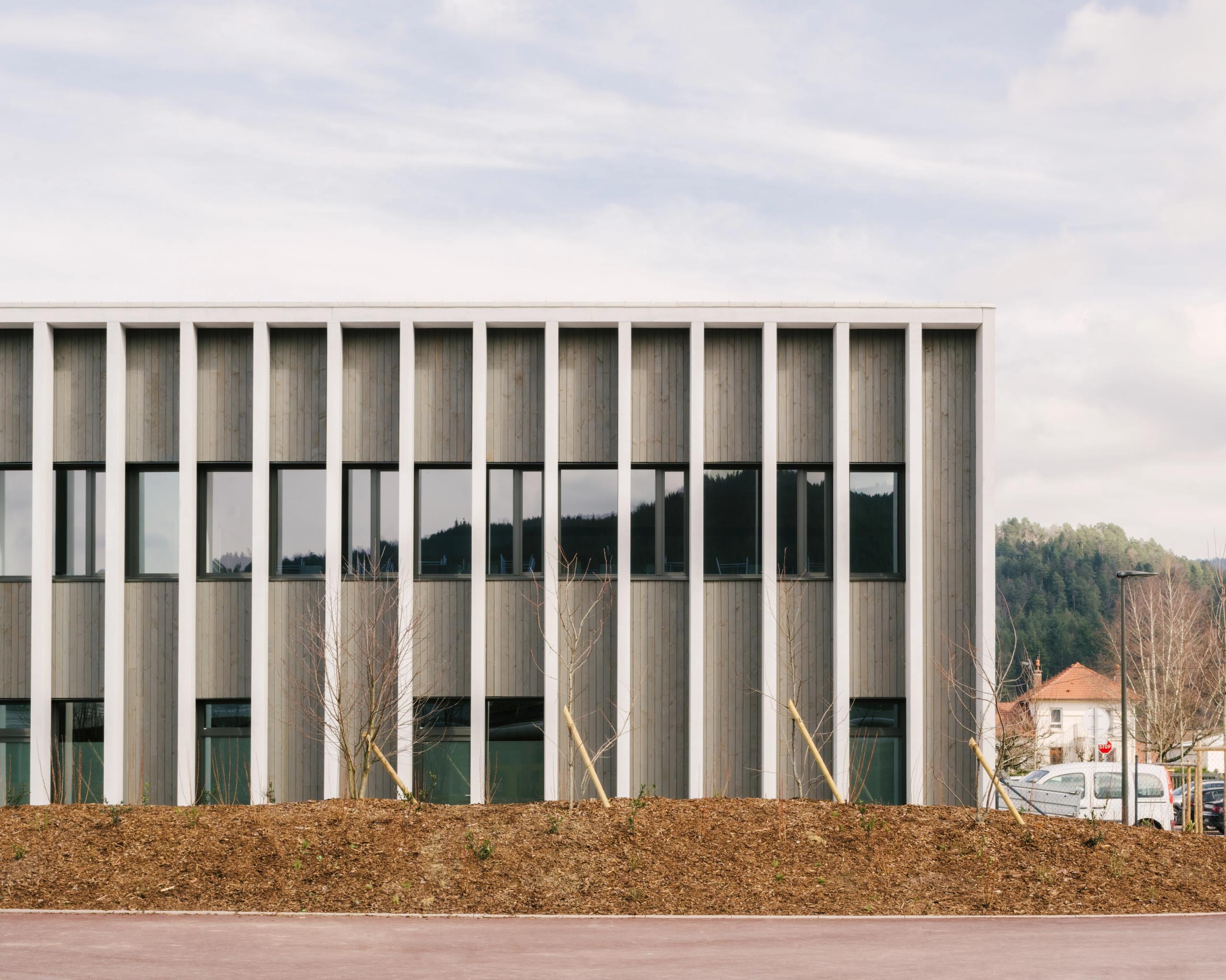 Façade bois-béton rythmée de poteaux verticaux et baies vitrées du Collège du Ban à Vagney, sur fond de paysage vosgien et d’aménagements neufs.