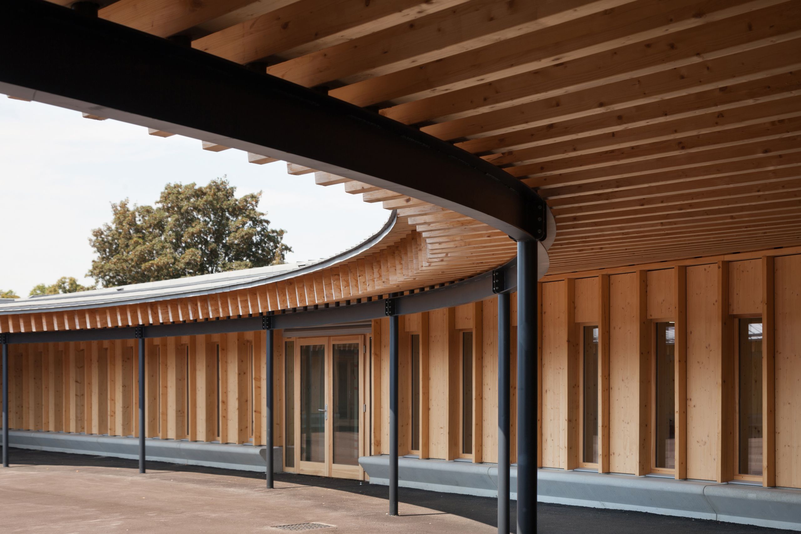 Cour couverte de l’école élémentaire Hubert Reeves à Champagnole, montrant la toiture elliptique en impluvium, l’ossature bois apparente et les poteaux métalliques.
