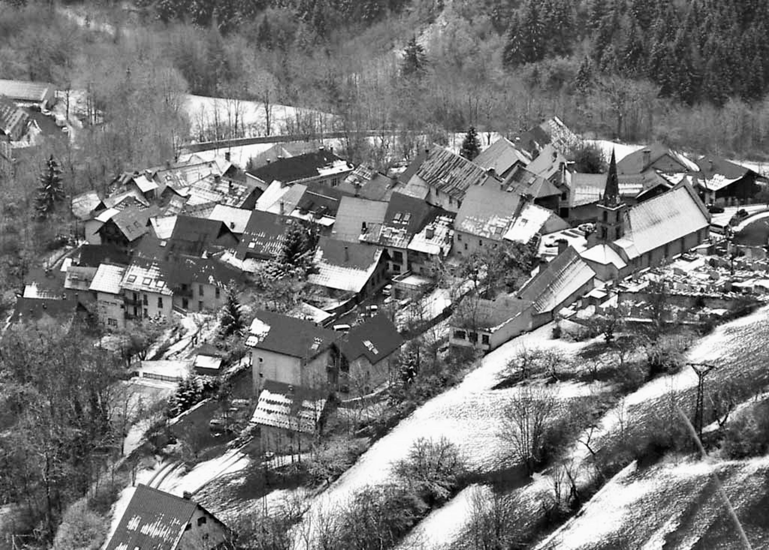 Vue aérienne en hiver du village des Deux Alpes, toitures en pente et maçonneries serrées au pied de la forêt, contexte bâti de l’auberge de jeunesse.