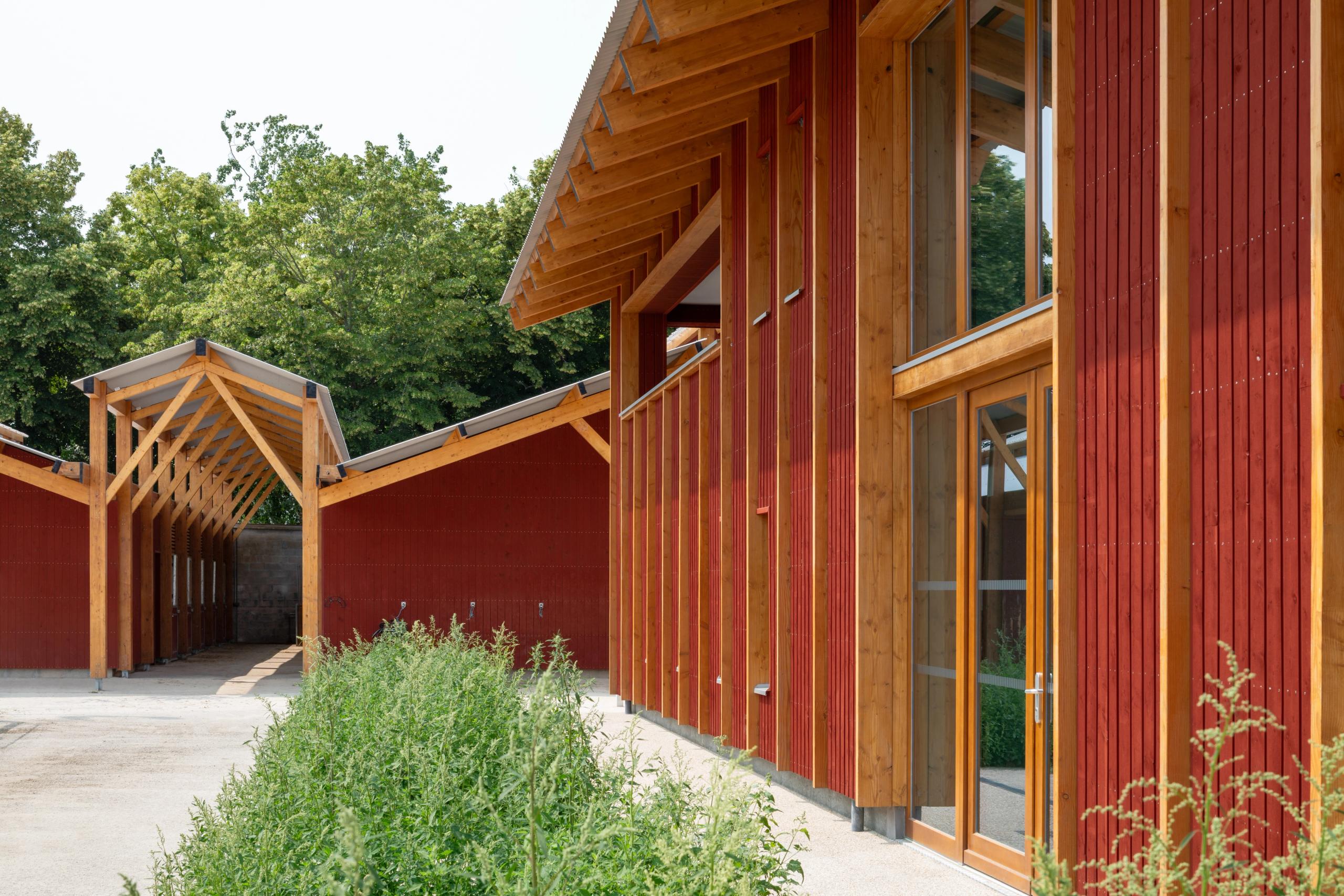 Timber-framed red-clad stables of the Gennevilliers equestrian center, with glazed openings and pitched roofs aligned along a planted courtyard.