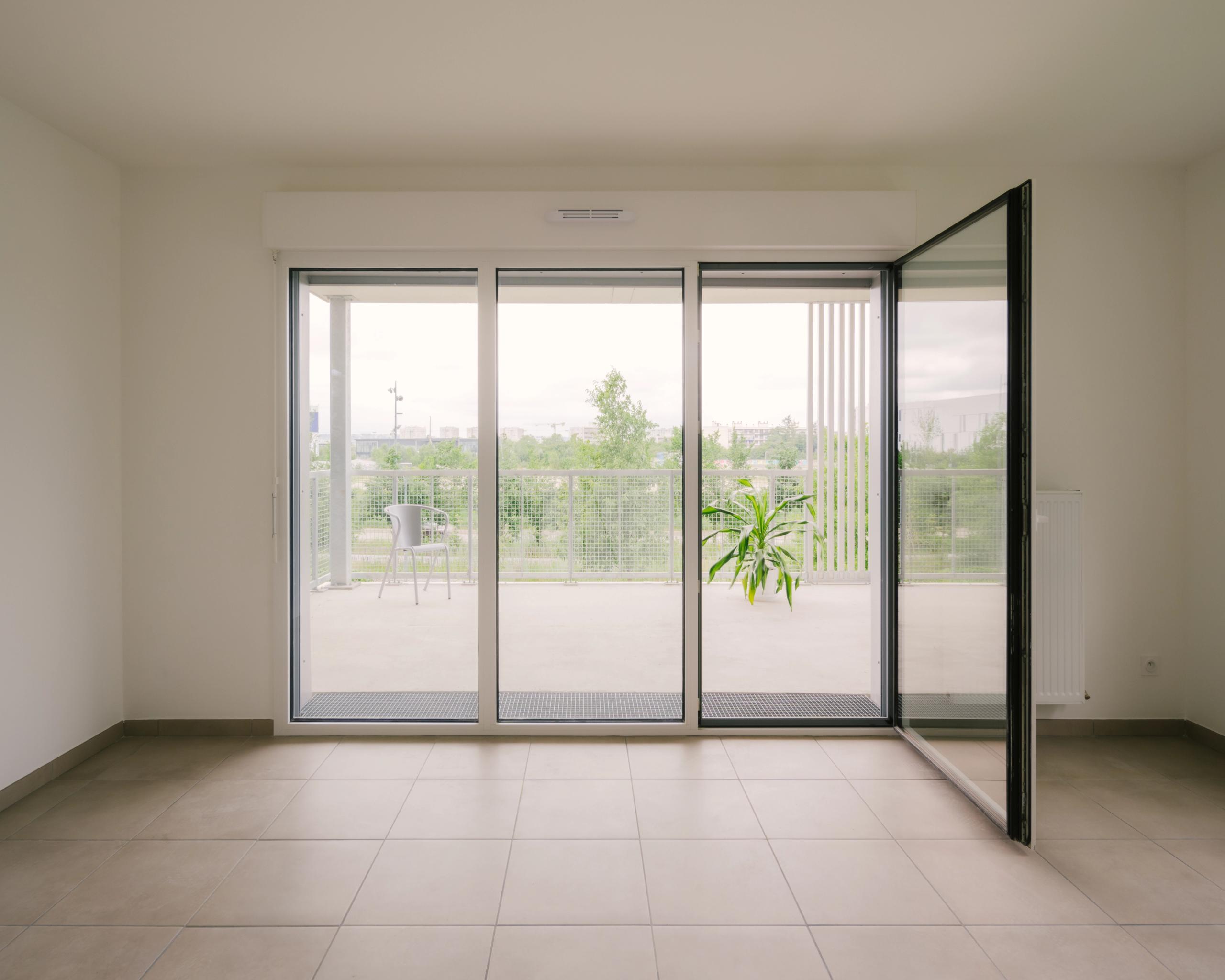 Intérieur d’un logement des 116 logements à Vénissieux, grande baie vitrée toute hauteur ouvrant sur balcon filant, garde-corps métal et vue paysagère.
