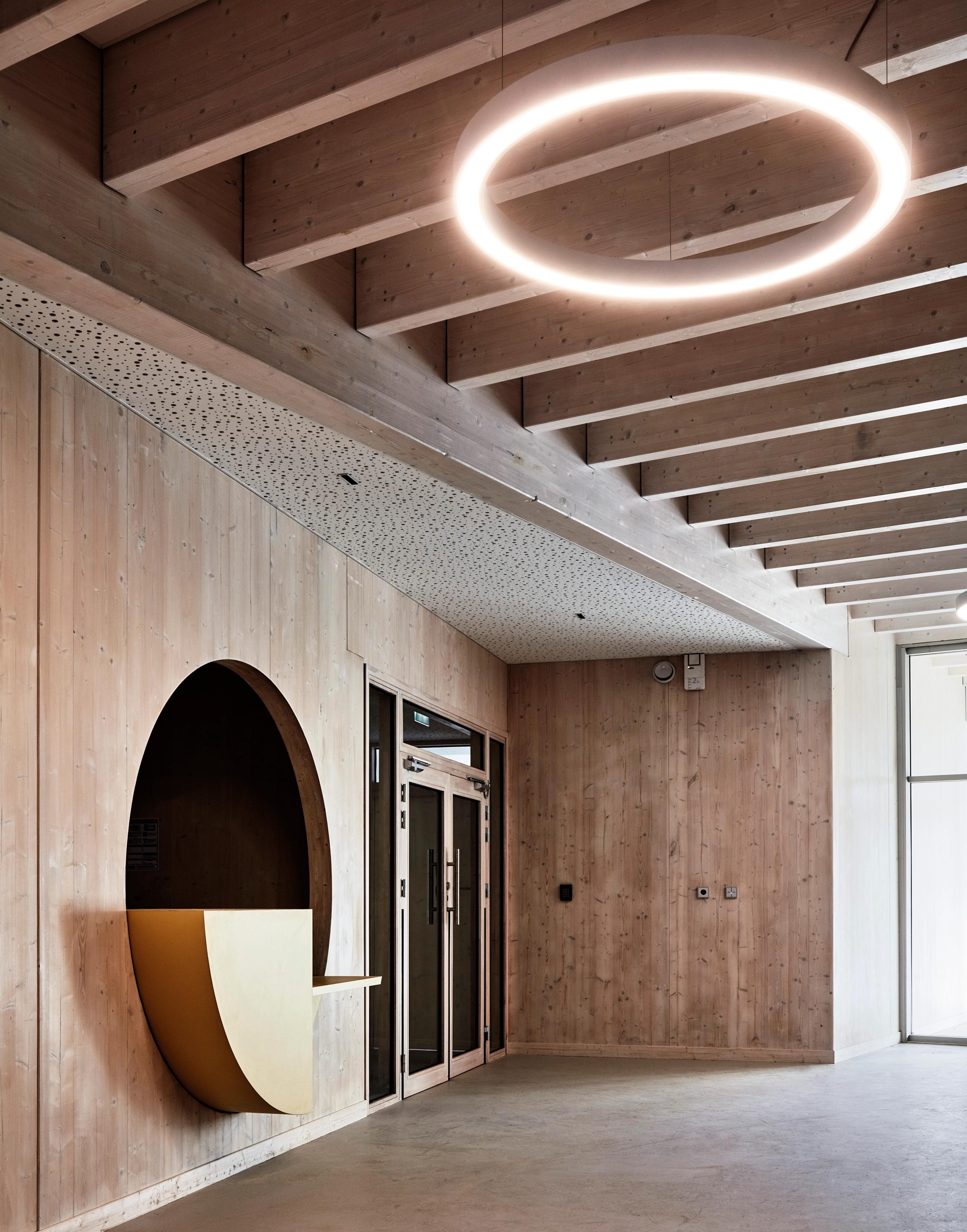 Hall de la Maison de l’Enfance Simone Veil à Albertville, structure bois apparente, comptoir circulaire jaune et luminaire annulaire suspendu.
