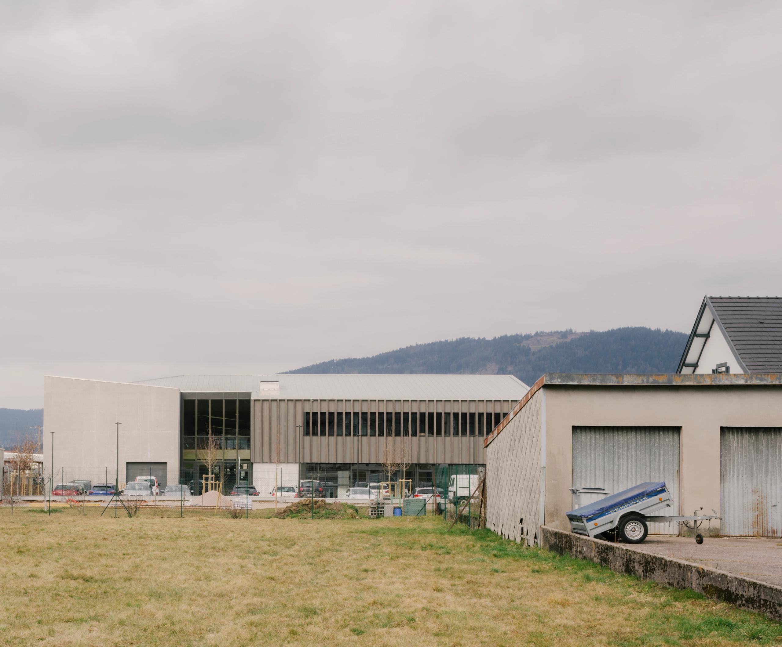Vue lointaine du Collège du Ban à Vagney : volumes en peigne bois-béton, façades rythmées de brise-soleil verticaux, implantés en plaine au pied du massif vosgien.