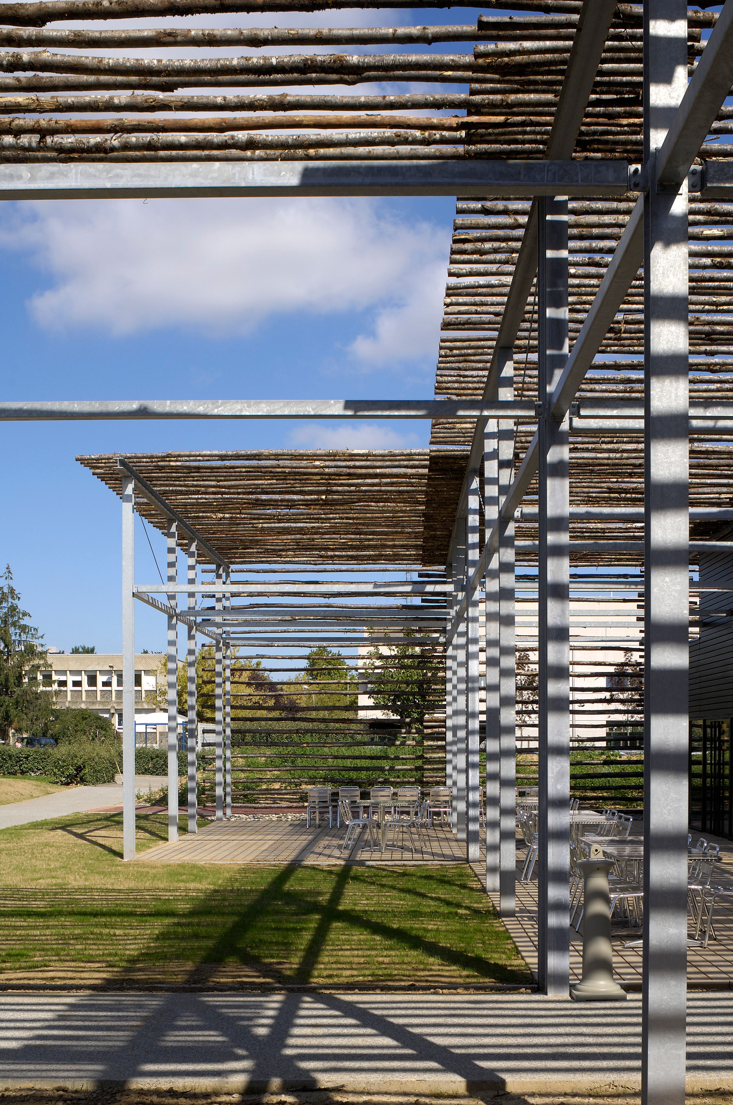 Terrasse du restaurant administratif du CETE à Bron, sous ombrière en baliveaux de châtaignier sur exostructure métallique, ombres portées et mobilier extérieur.