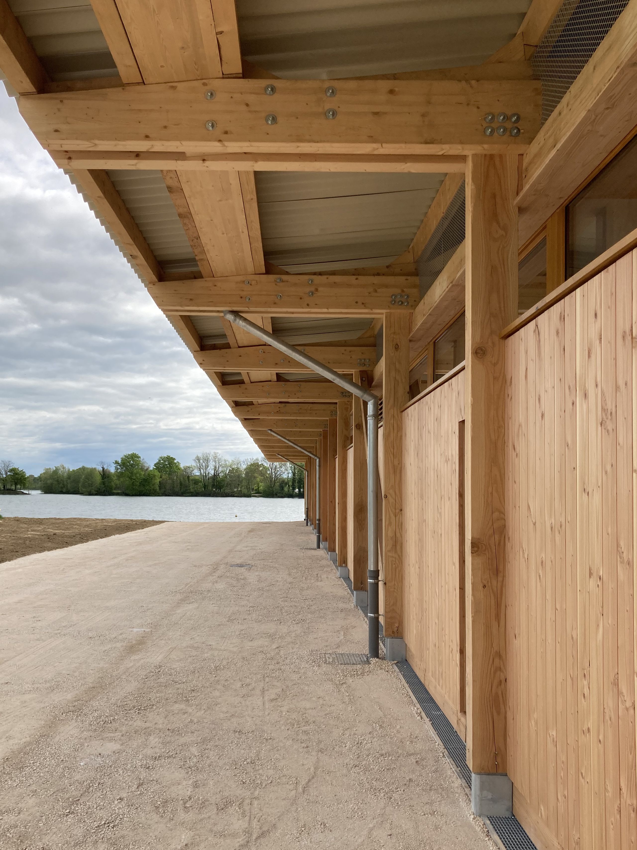 Façade en bois de la Maison des sports à Malafretaz, auvent structurel en ossature apparente, gouttières métalliques et promenade minérale face au plan d’eau.