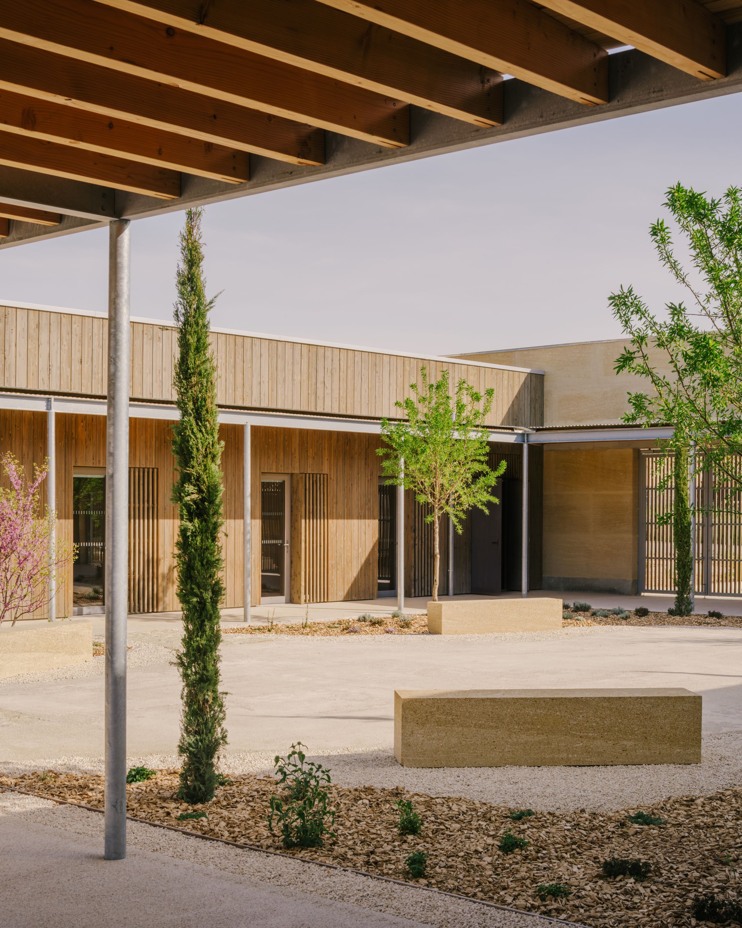 Timber and rammed-earth courtyard of the Youth Cultural Centre in Vaison-la-Romaine, with covered walkway, planted beds and monolithic seating blocks.