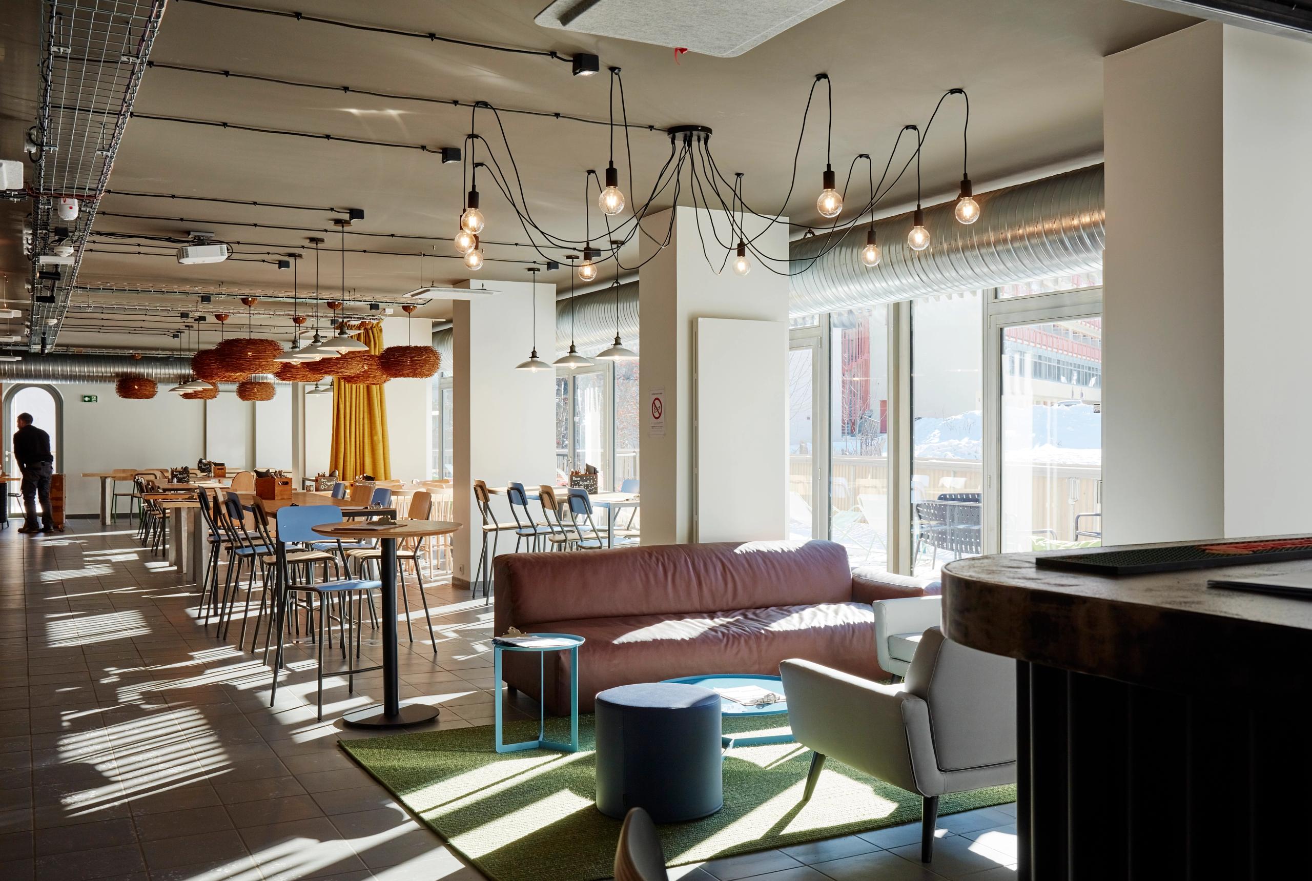 Espace commun de l’auberge de jeunesse des Deux Alpes par Tectoniques, vaste plateau vitré avec mobilier léger, luminaires apparents et plafond technique.