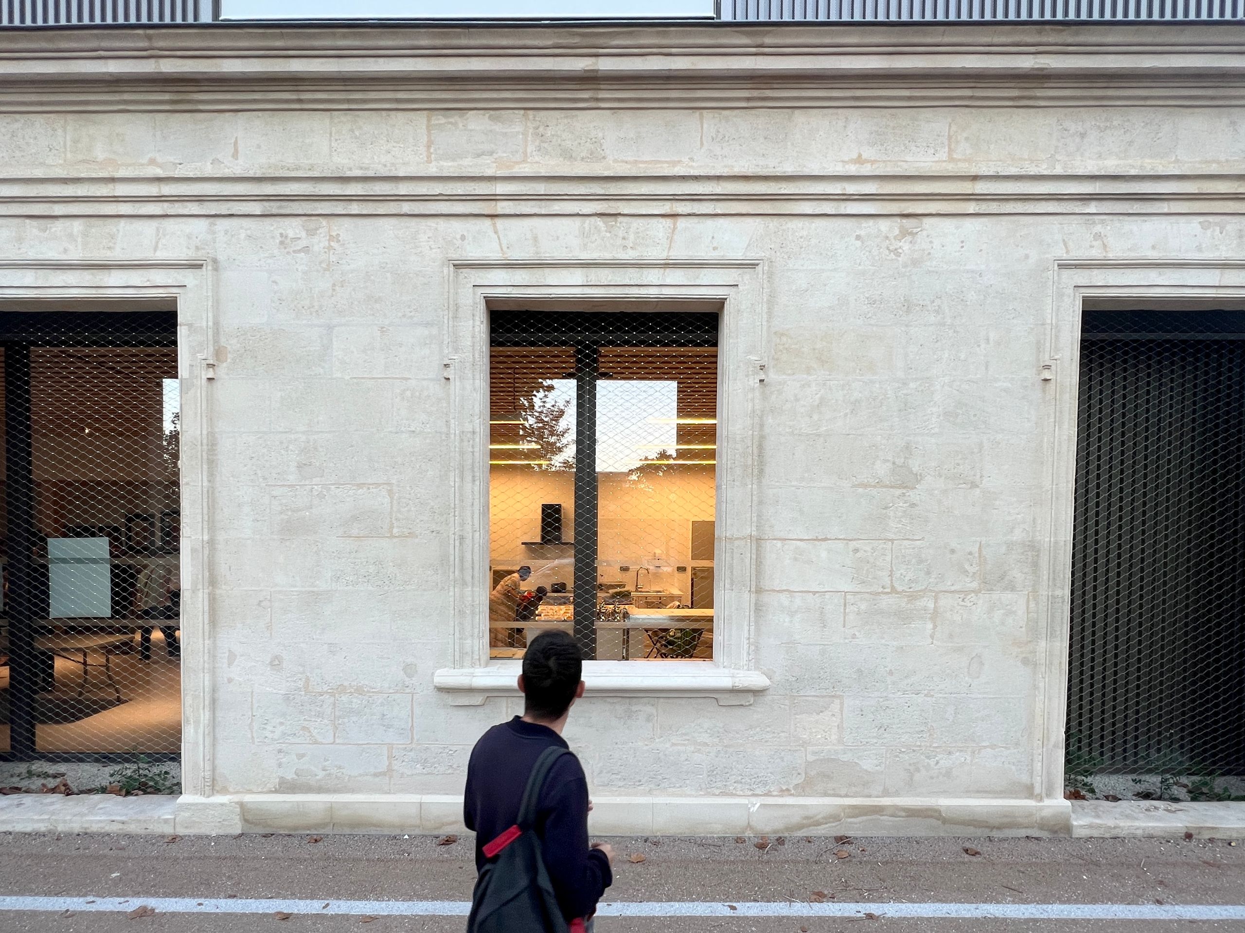Façade rénovée de la maison des habitants à Mérignac, parement en pierre claire, baies protégées par treillis métallique, vue intérieure sur espace d’accueil éclairé.