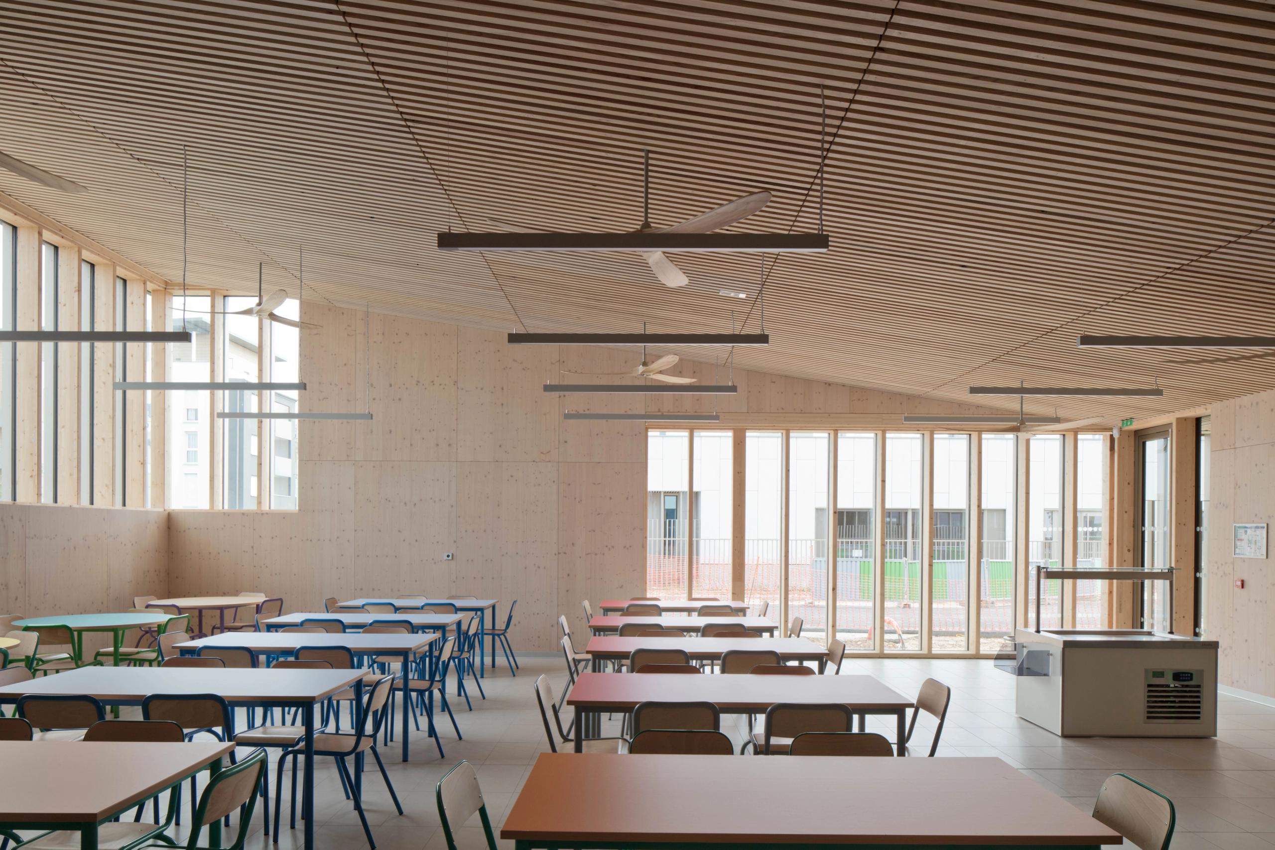 Cafeteria du groupe scolaire Denis Mukwege à Melun, vaste volume en bois avec plafond à lames, grandes baies sud et mobilier coloré organisé en trames régulières.