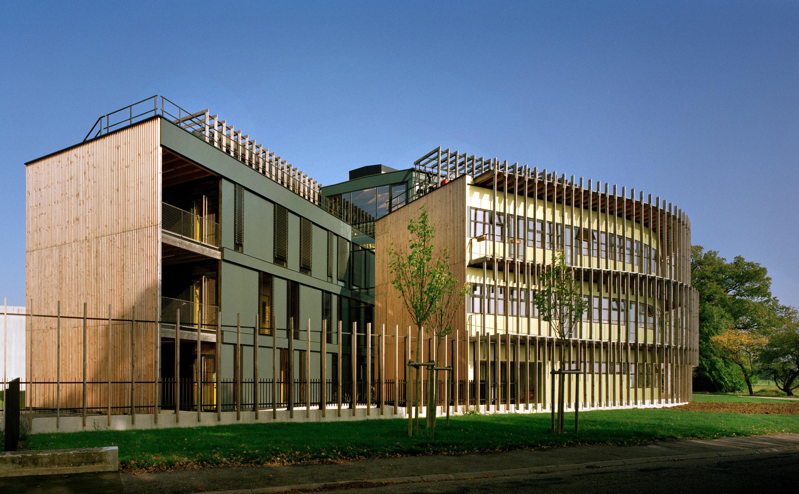 Vue du laboratoire INRAE à Champenoux : deux ailes bois-béton reliées par un atrium vitré, façade sud courbe protégée par un brise-soleil vertical en bois.