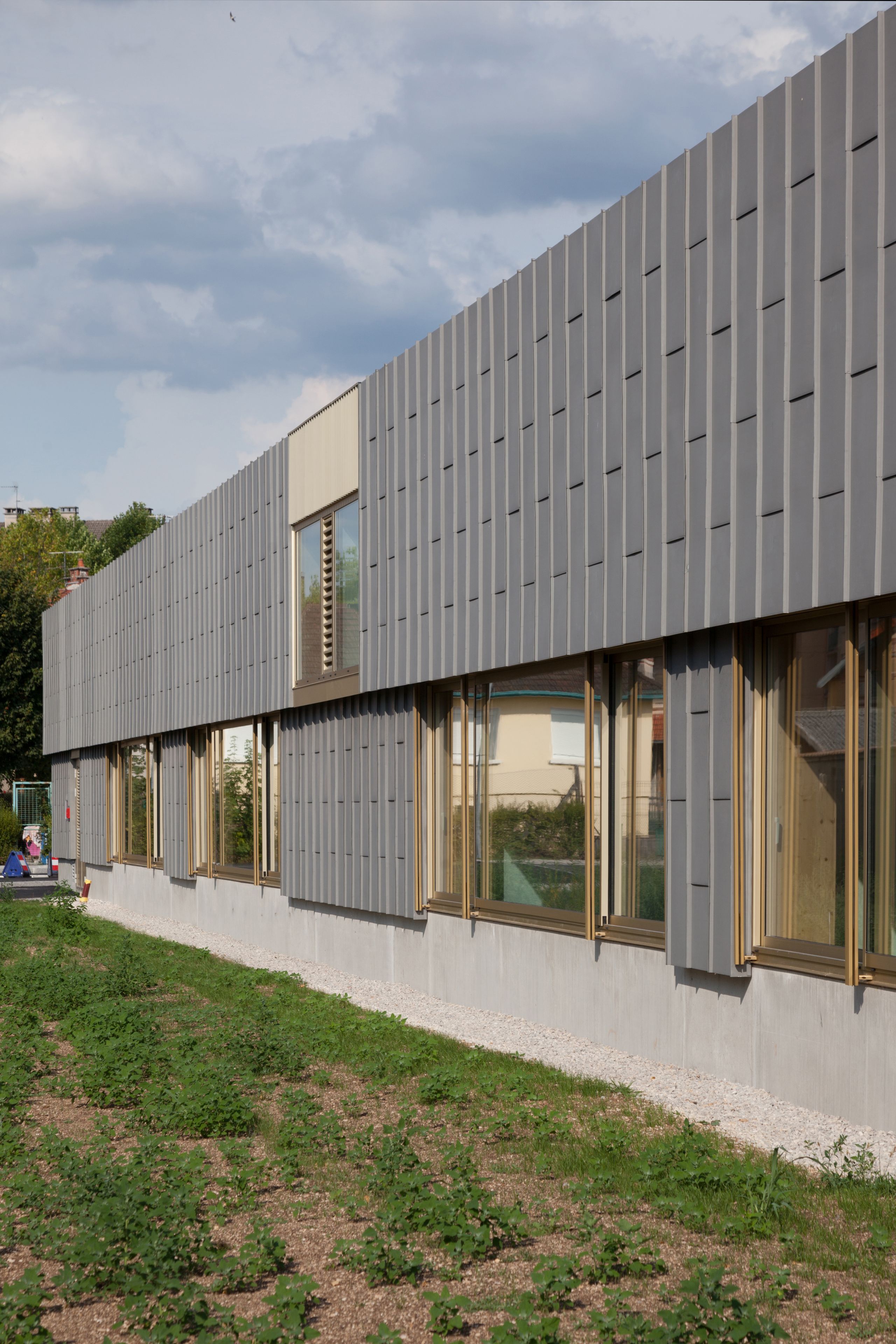 Façade de l’école élémentaire Hubert Reeves à Champagnole, bardage zinc rythmé et larges baies bois vitrées ouvrant sur un jardin en lisière périurbaine.