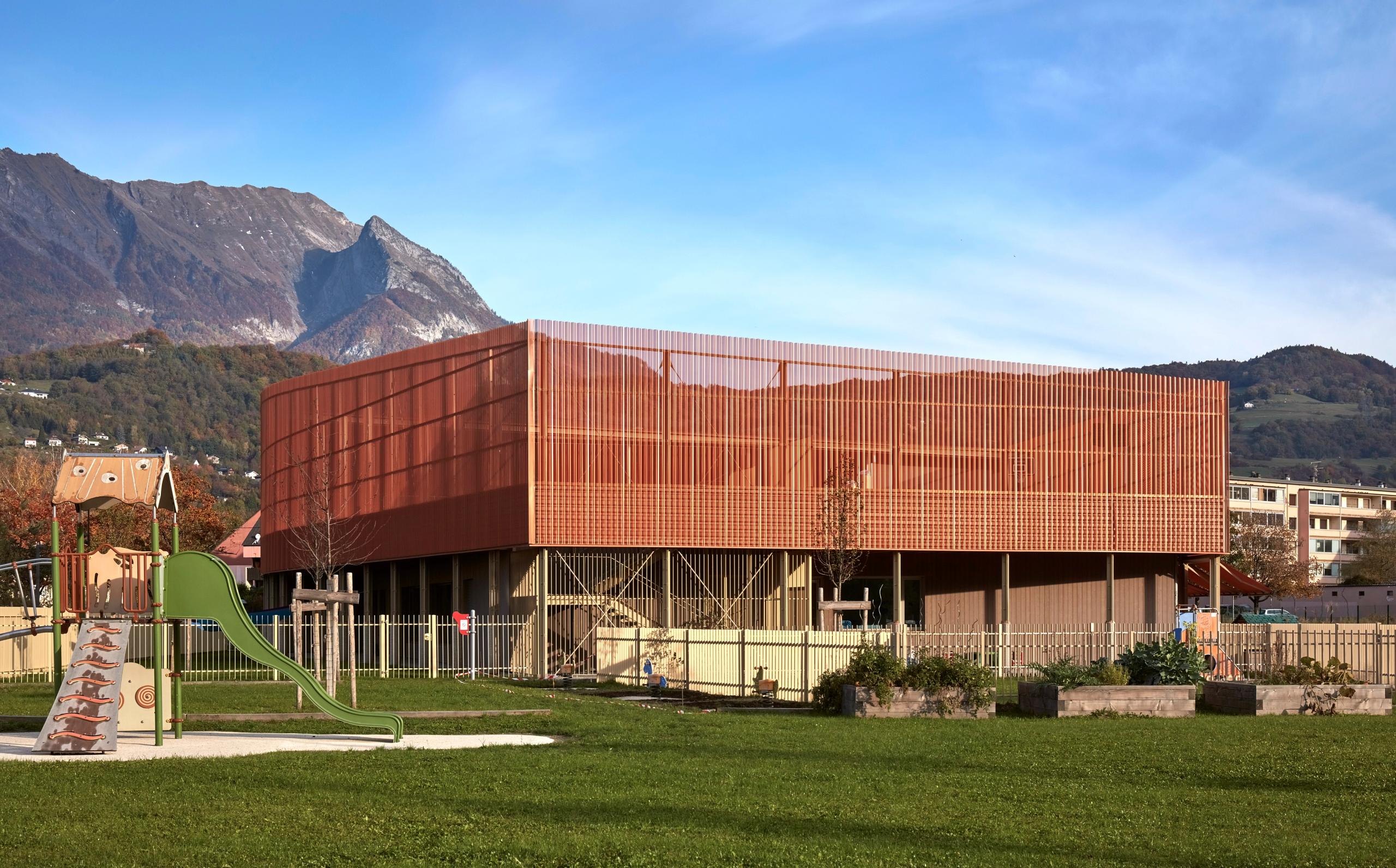Maison de l’enfance Simone Veil à Albertville, volume compact en métal cuivré sur socle bois-béton, surélevé sur pilotis face aux massifs alpins.