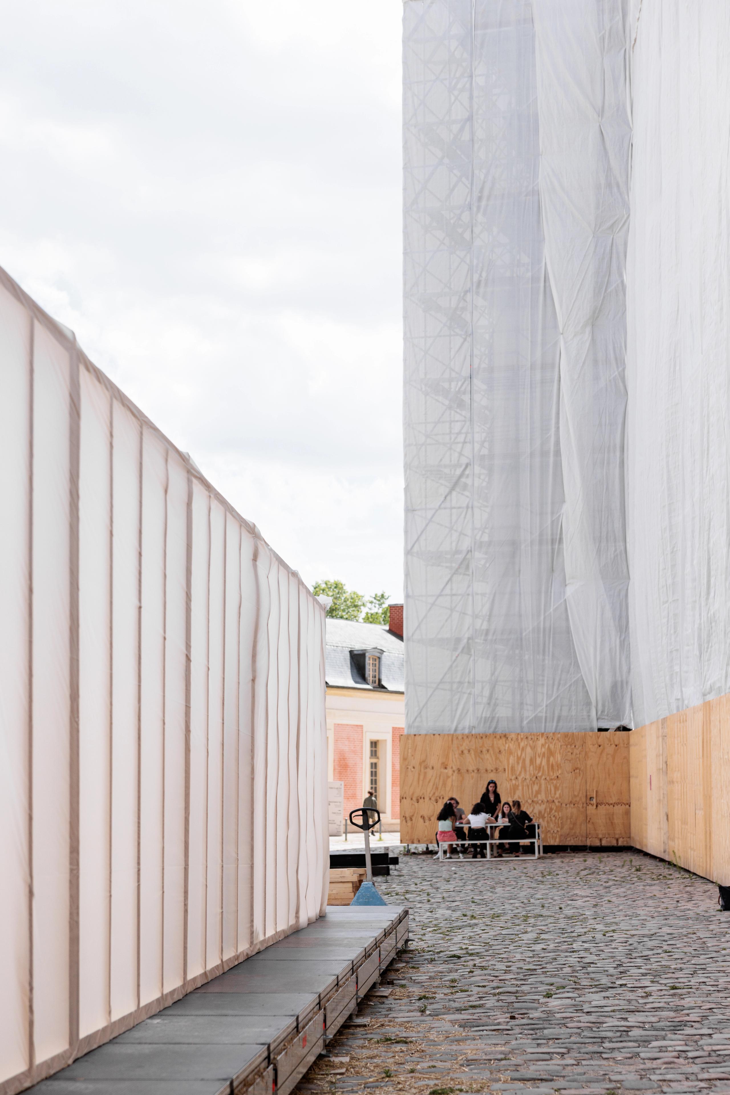 Workshop 60x60 à Versailles : couloir extérieur entre parois bois et film plastique translucide, sol pavé, groupe assis au fond de la Cour de la Maréchalerie.